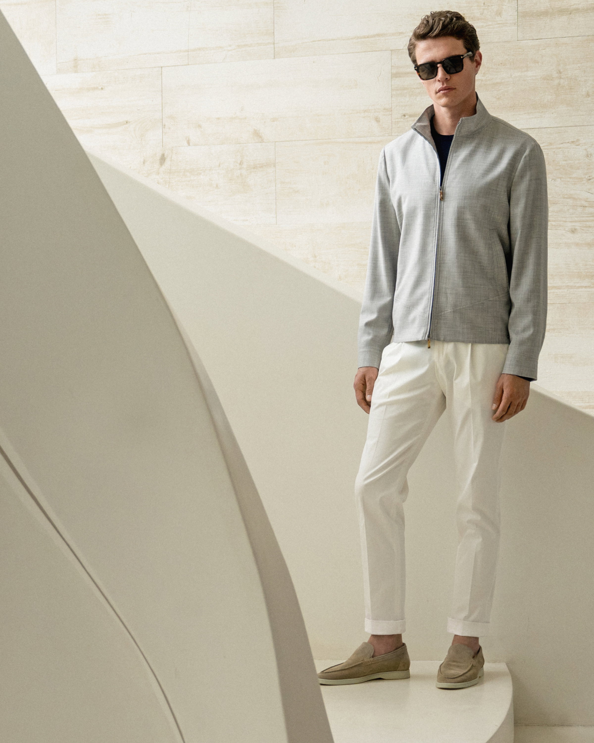 Mann steht auf weißer Treppe, trägt hellgraue Jacke, weißer Hose, beige Loafer und schwarze Sonnenbrille vor hellem Hintergrund.
