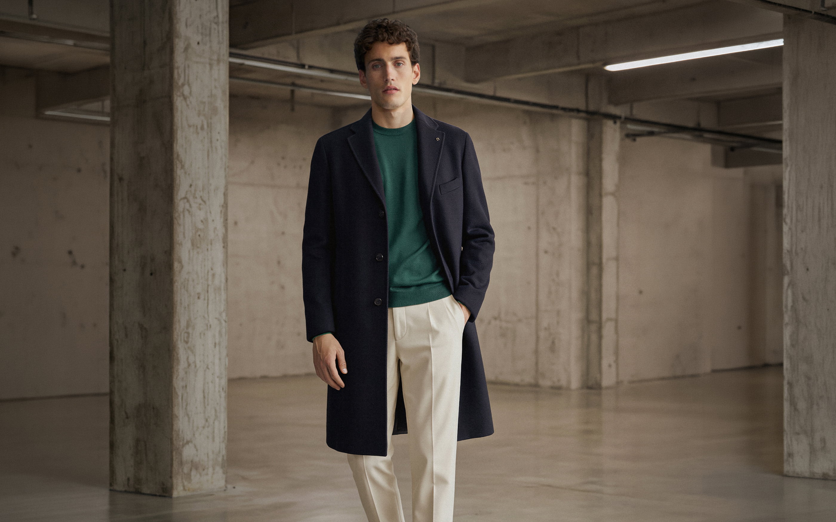 Mann in cremefarbener Hose, grünem Pullover und dunkelblauem Wollmantel steht in einem leeren Industriegebäude mit Betonstützen.