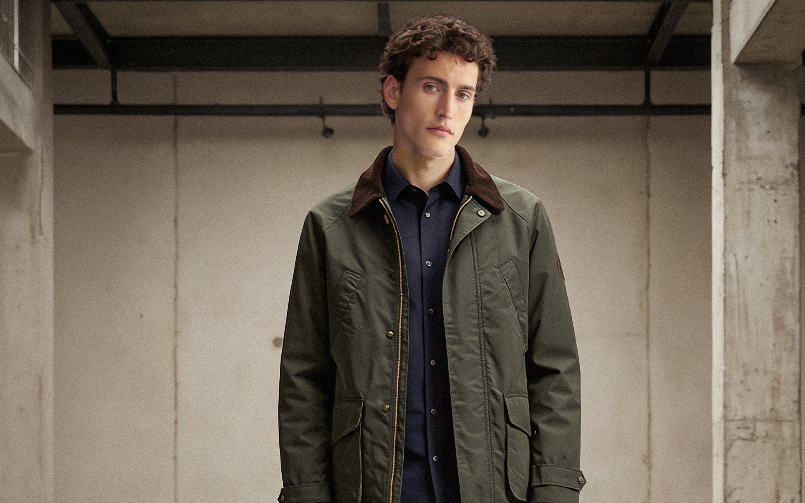 Junger Mann mit lockigem Haar trägt olivgrünen Parka mit braunem Kragen und dunkelblaues Hemd in Industriegebäude.