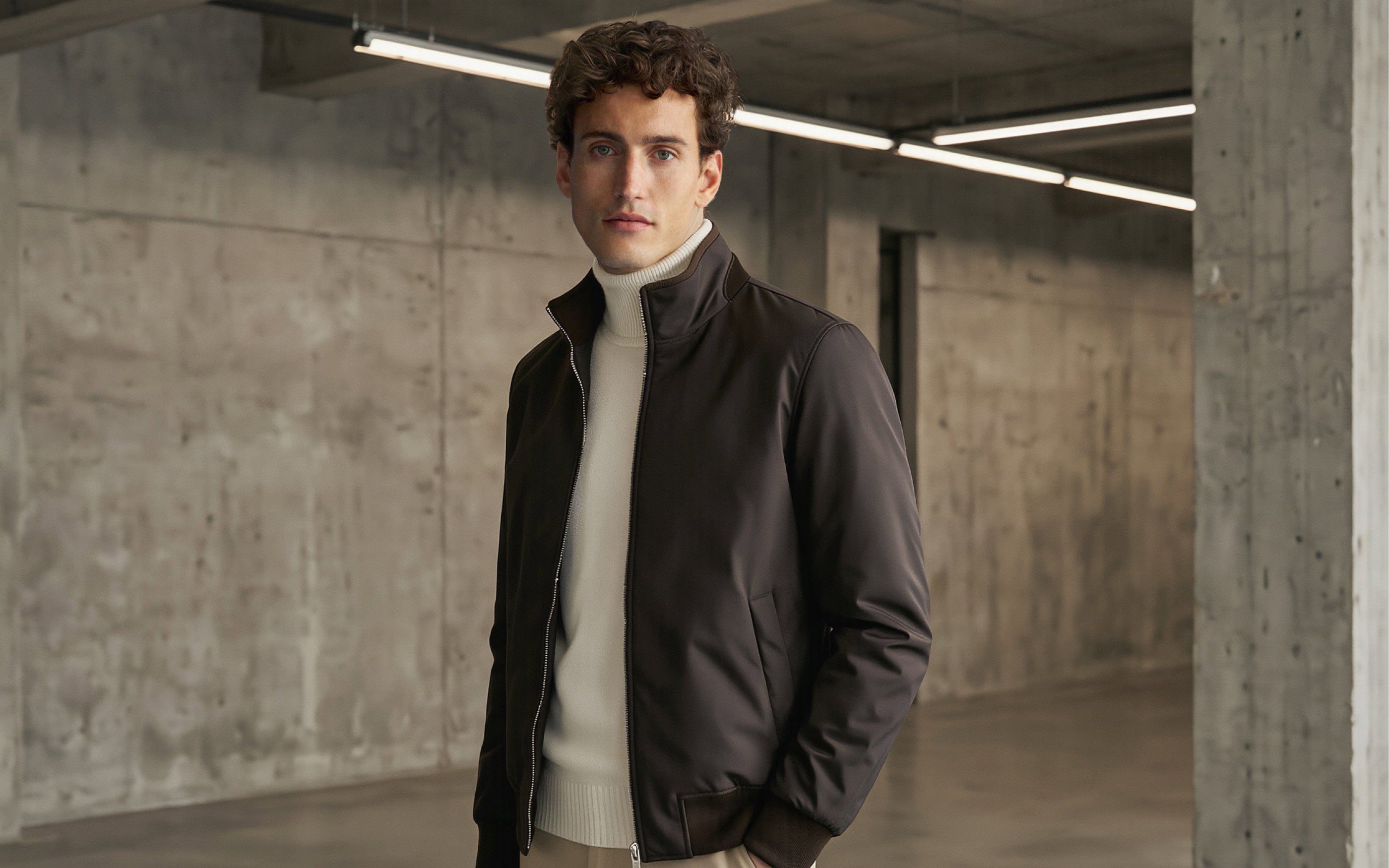 Junger Mann mit weißem Rollkragenpullover und dunkelbraunem Blouson in einem kargen Industriegebäude mit Betonwänden.