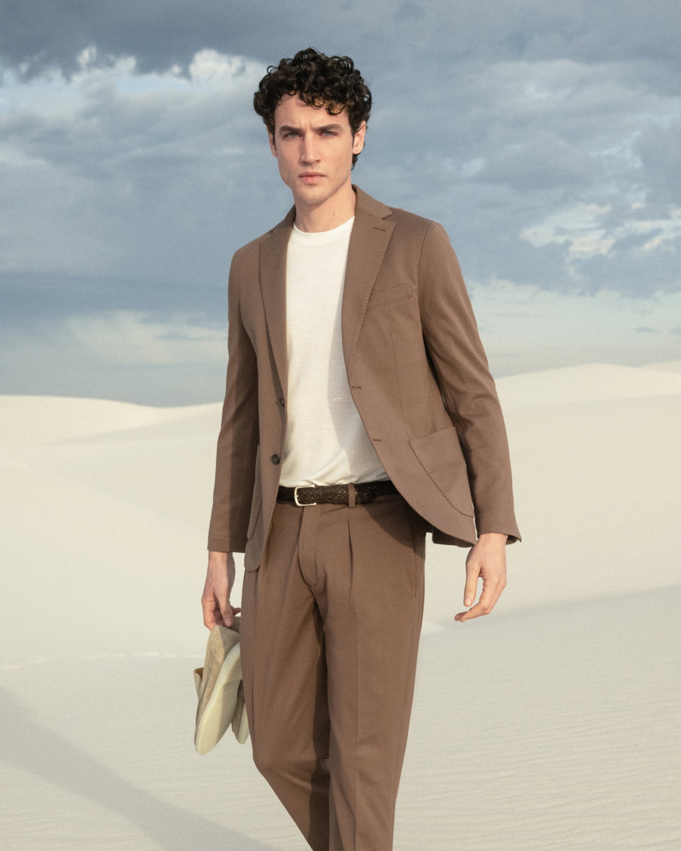 Mann mit braunem Anzug und weißem Shirt läuft durch helle Sanddünen, trägt Sneaker in der Hand, bewölkter Himmel.