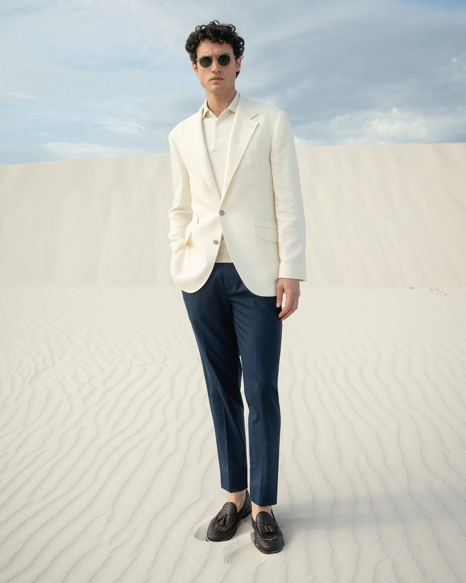 Mann in Sanddünen trägt cremefarbenes Poloshirt und Sakko, navy Hose, braune Loafer und runde Sonnenbrille.