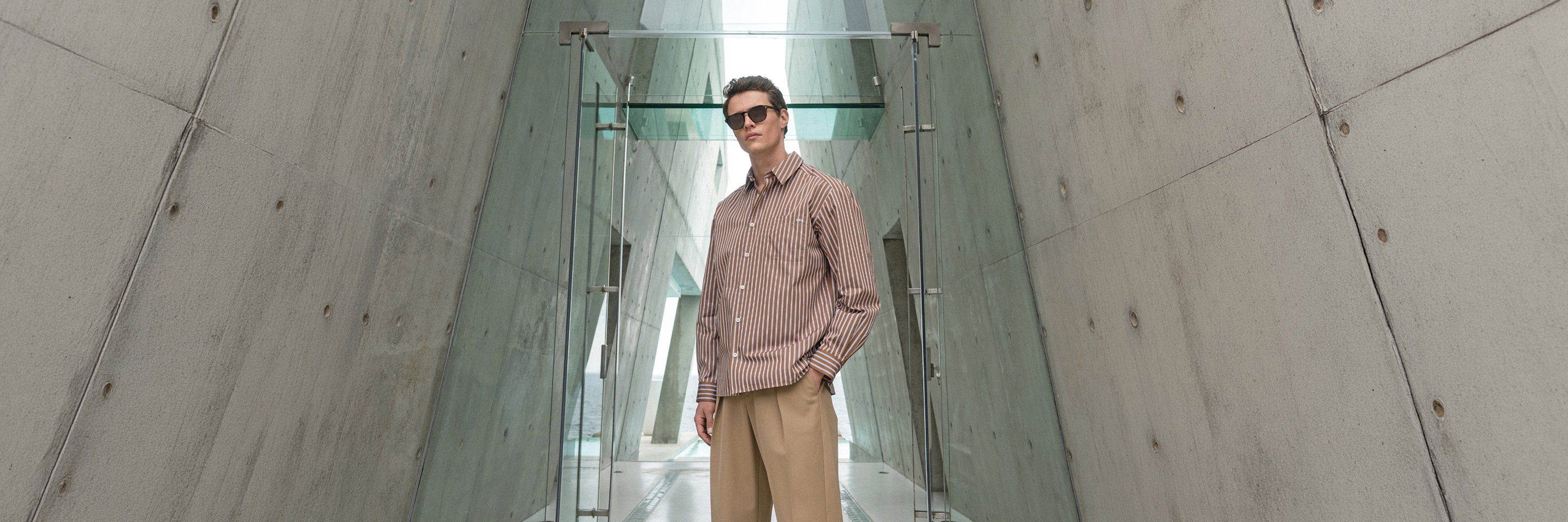 Mann mit beiger Hose, gestreiftem Hemd und Sonnenbrille steht vor Glastür in modernem, betonverkleidetem Gebäude.