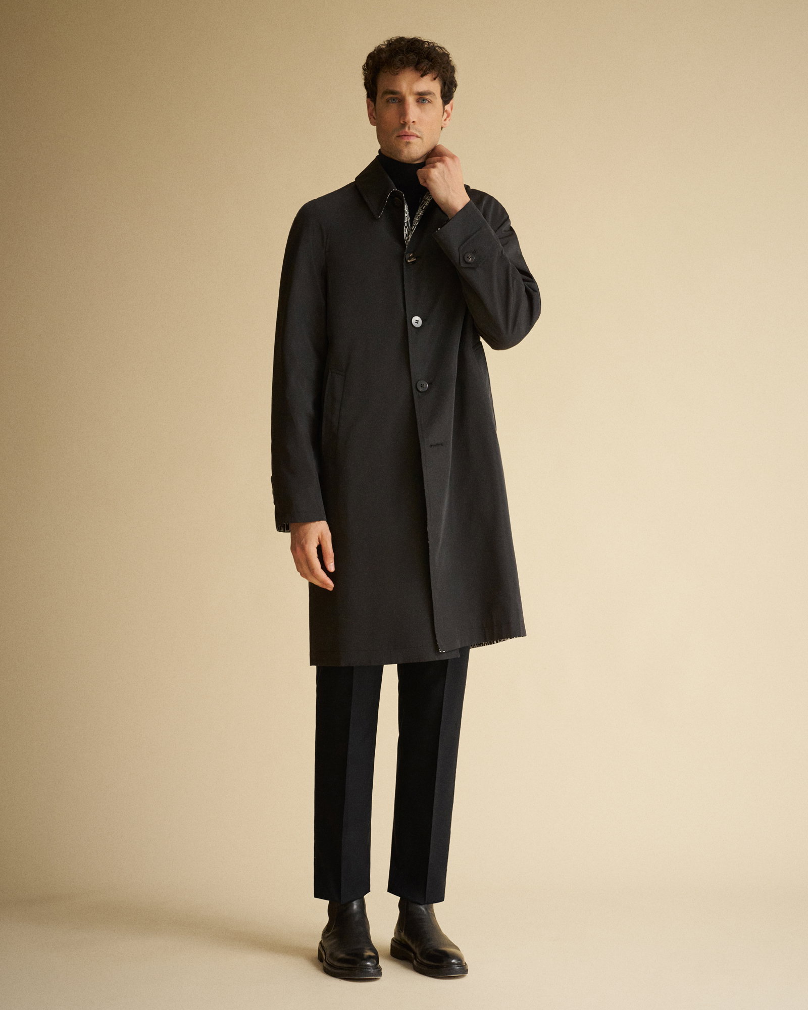 Mann mit schwarzen Lederboots, schwarzer Hose, schwarzem Rollkragenpullover und langem schwarzem Wollmantel vor beigem Hintergrund.