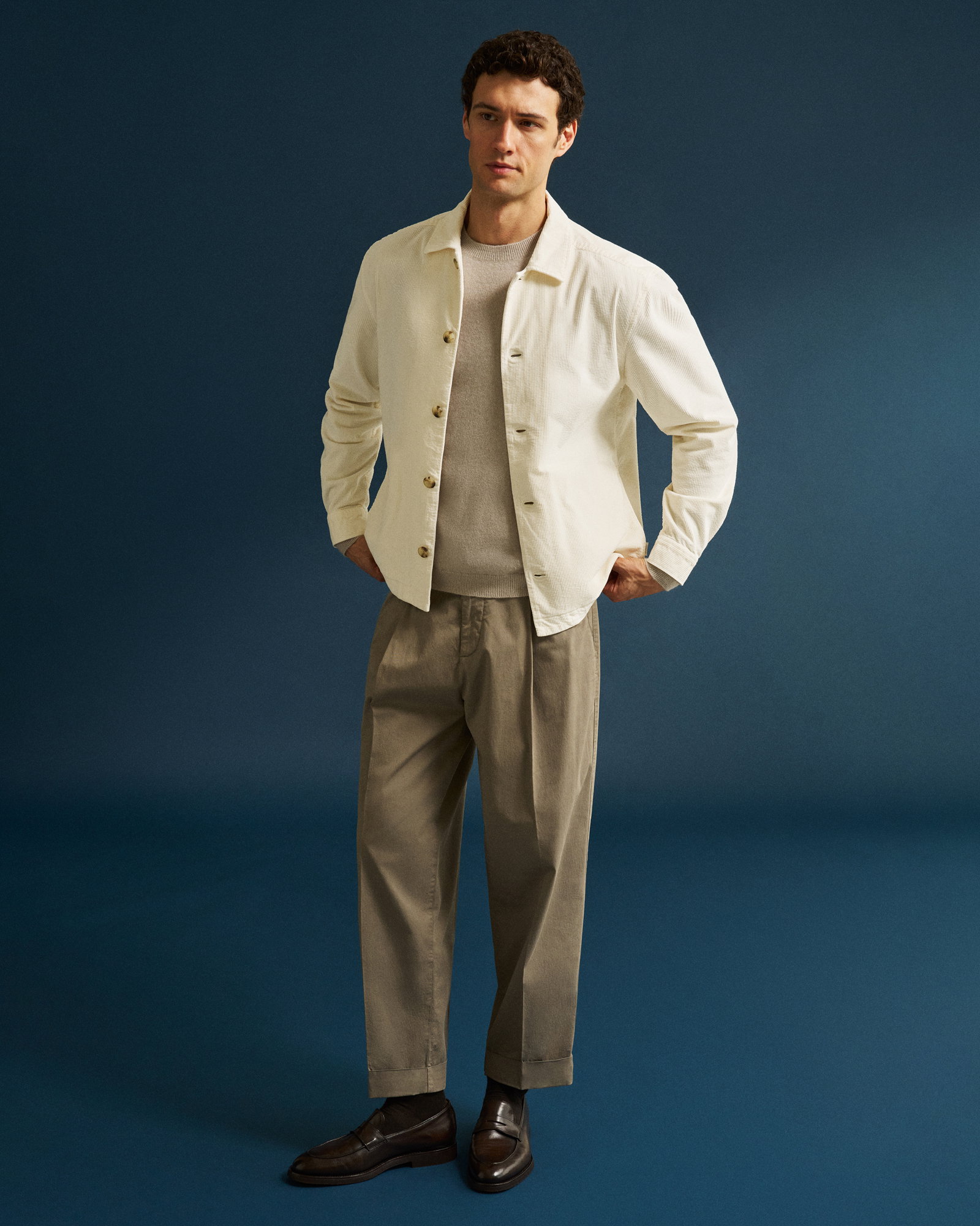 Mann trägt cremefarbenes Overshirt, beigen Rundhalspullover, khakifarbene Chinohose und dunkelbraune Loafer vor blauem Hintergrund.