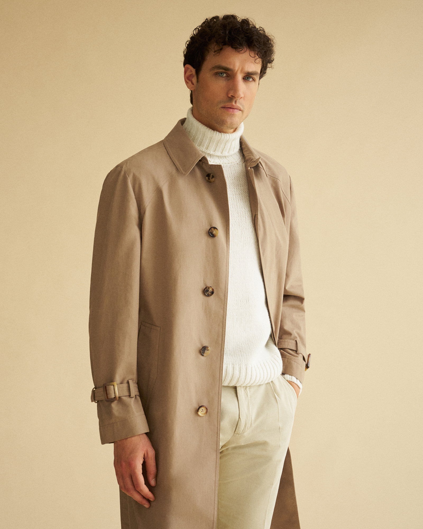 Mann mit dunklem lockigem Haar trägt beigefarbenen Trenchcoat, cremefarbenen Rollkragenpullover und helle Hose vor sandfarbenem Hintergrund.