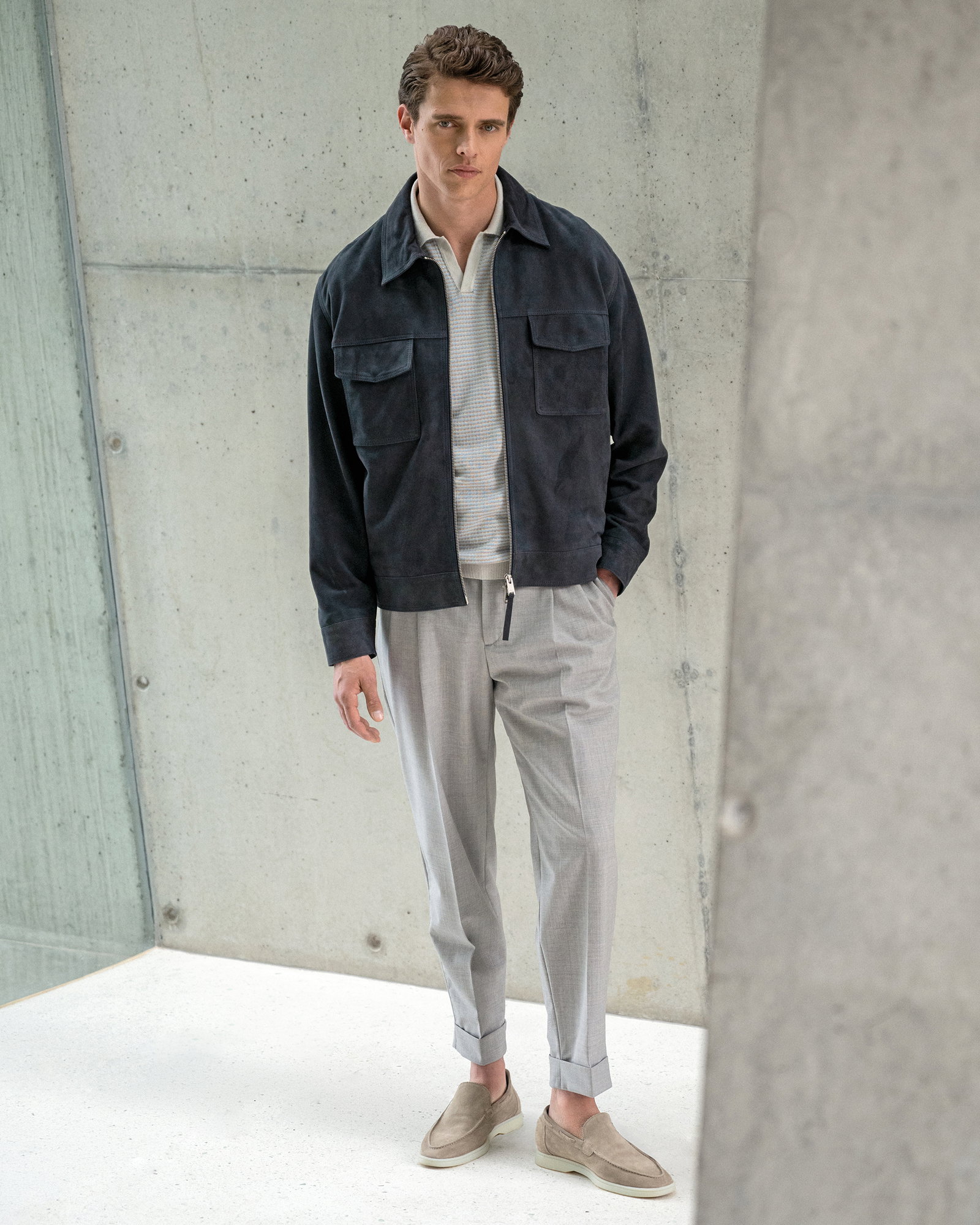 Mann trägt navy Velourslederjacke, graues Strickpolo, hellgraue Hose und beige Loafer vor Betonwand stehend.