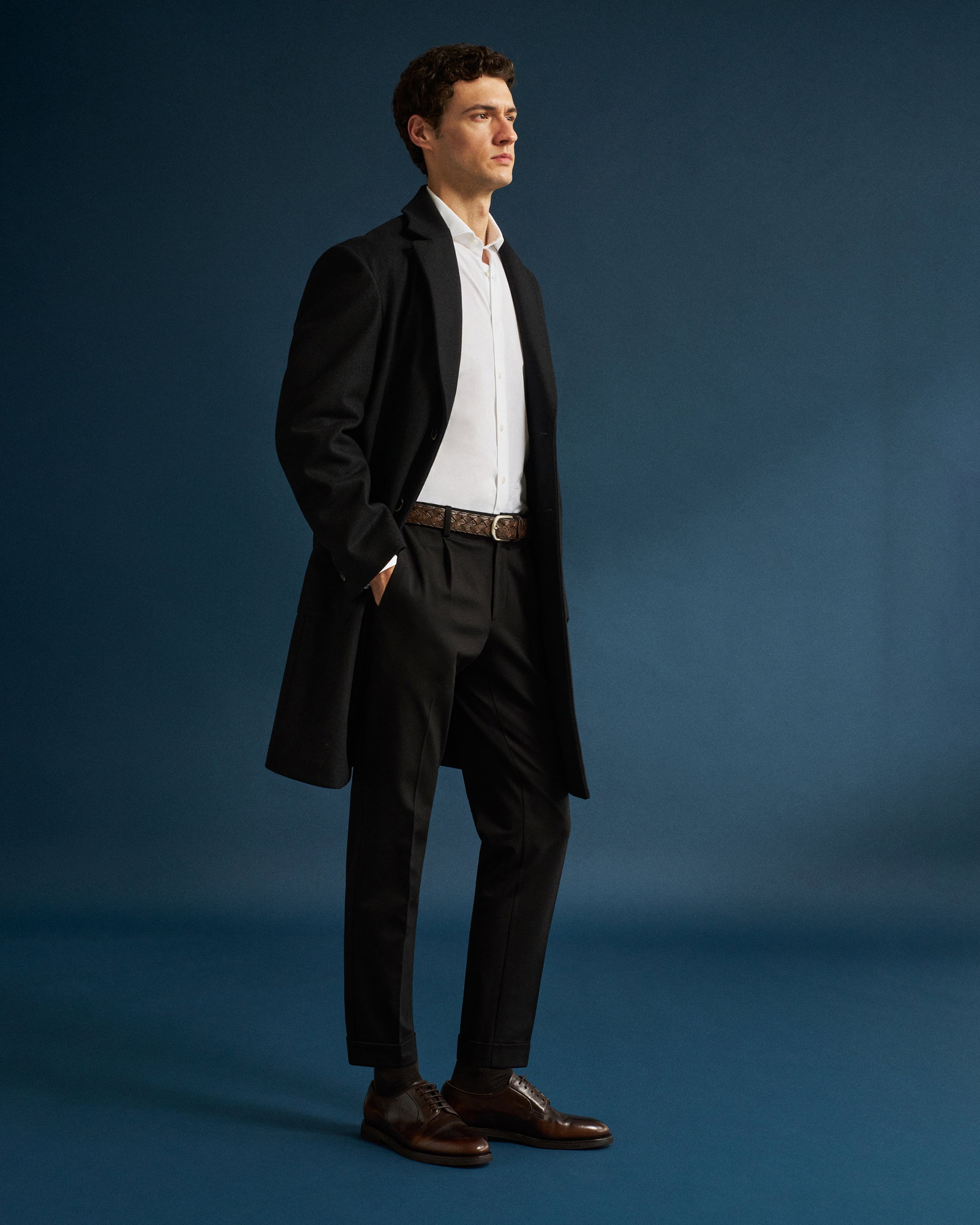 Mann steht vor blauem Hintergrund, trägt schwarzen Wollmantel, weißes Hemd, schwarze Hose, braunen Gürtel und braune Schnürschuhe.