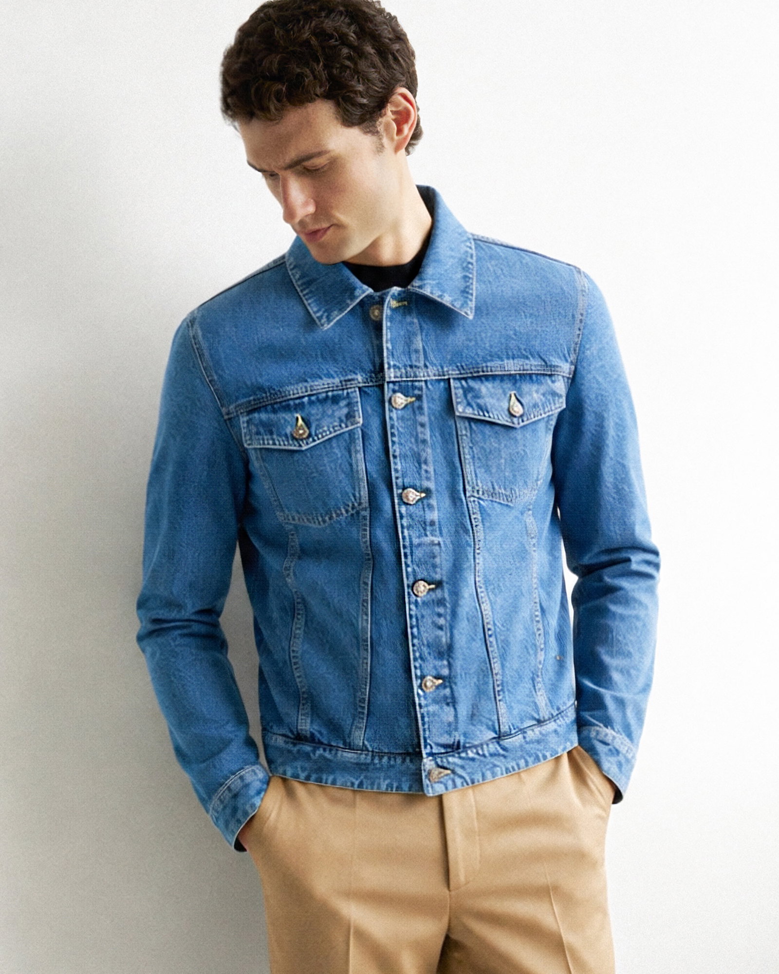 Mann mit kurzen dunklen lockigen Haaren trägt blaue Jeansjacke, schwarzes Shirt und beigen Hose, schaut nach unten.