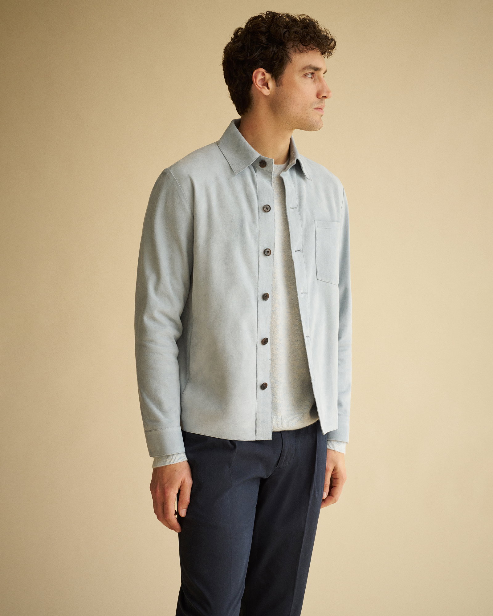 Mann mit lockigen dunklen Haaren trägt hellblaues Overshirt, hellblauen Pullover und dunkelblaue Hose vor sandfarbenem Hintergrund.