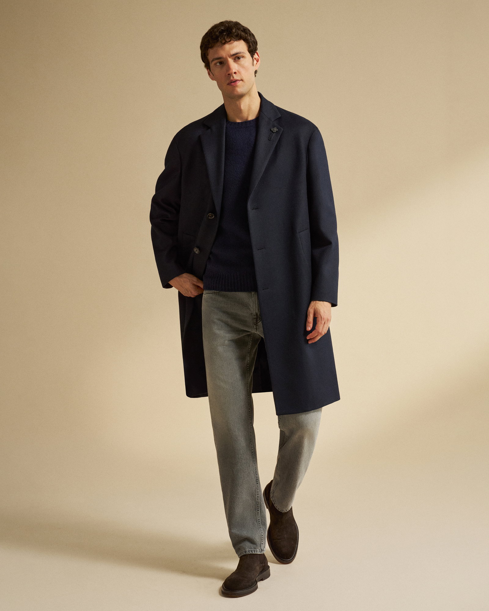 Mann mit kurzen dunklen Haaren trägt dunkelblauen Mantel, blauen Pullover, graue Jeans und braune Chelsea-Boots vor beigem Hintergrund.