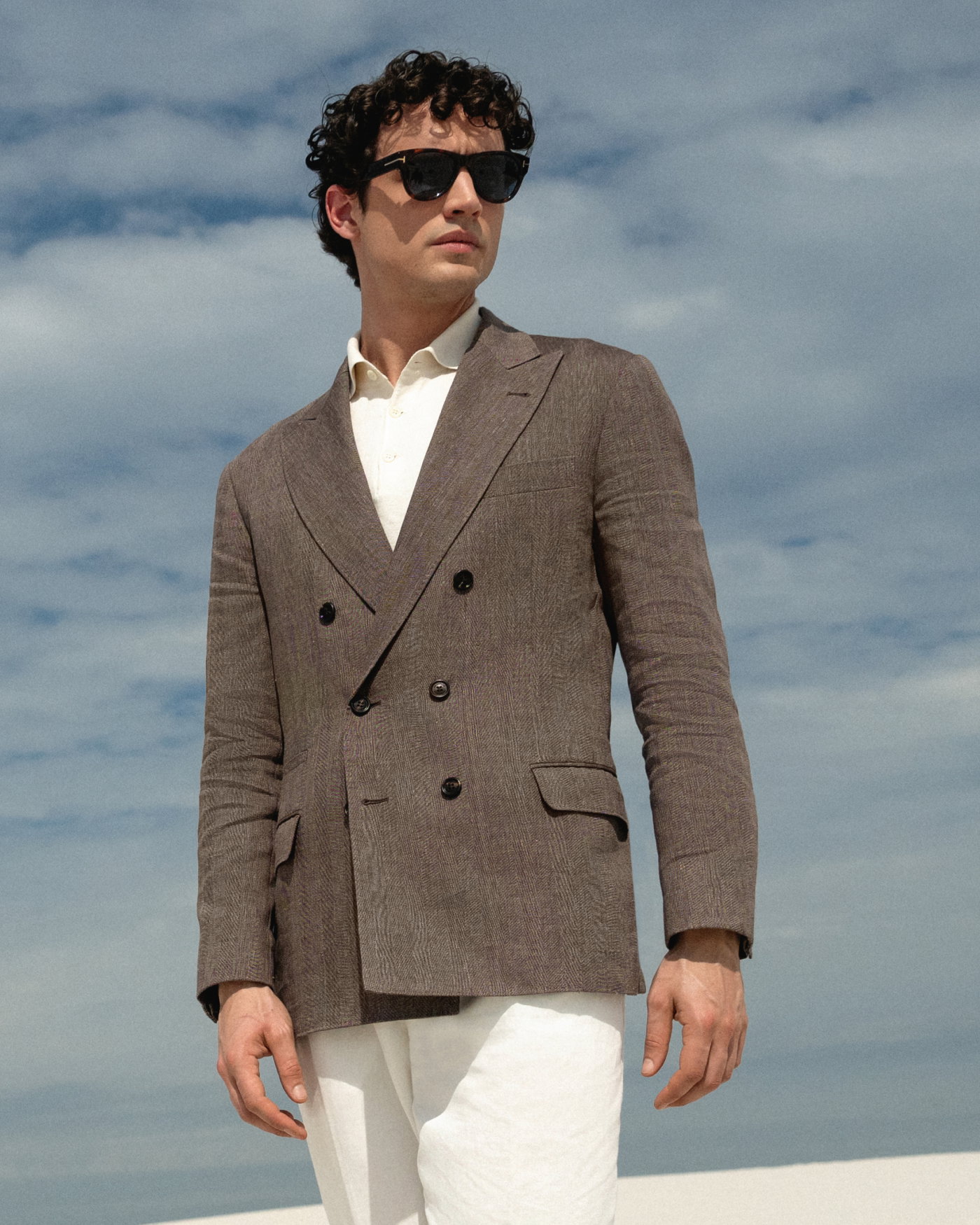 Mann mit lockigem Haar trägt dunkelbraunes Sakko, weißes Poloshirt, weiße Hose und schwarze Sonnenbrille vor bewölktem Himmel.