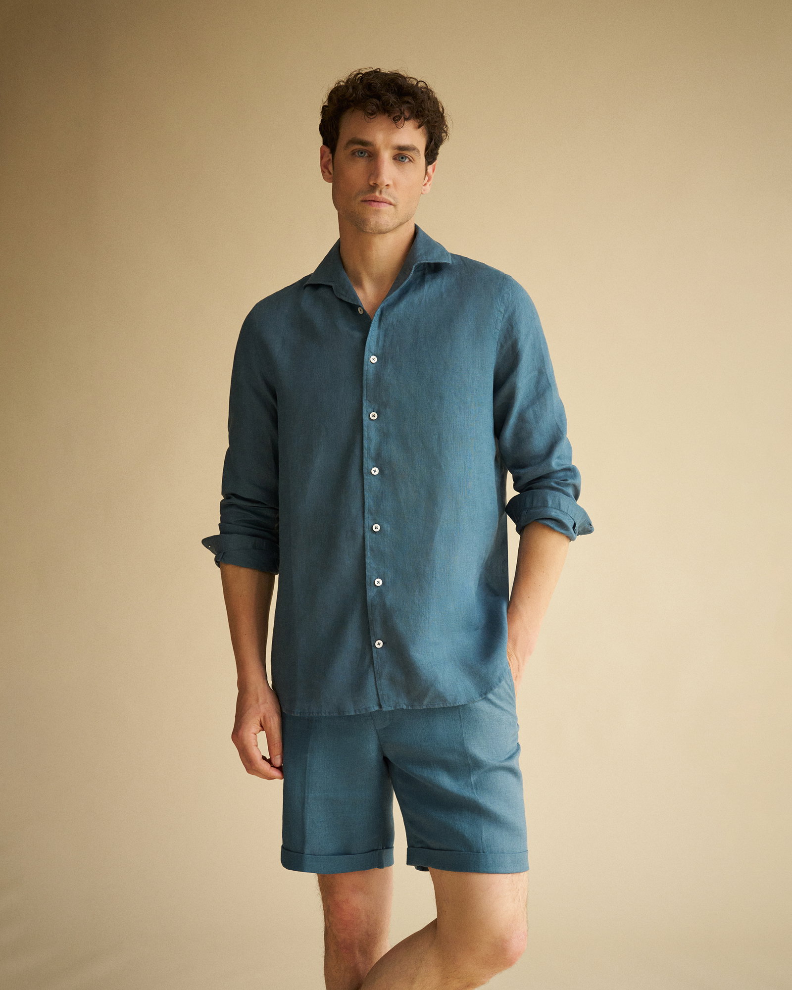 Mann mit lockigem Haar trägt blaues Leinenhemd und passende Shorts, steht vor hellem einfarbigem Hintergrund.