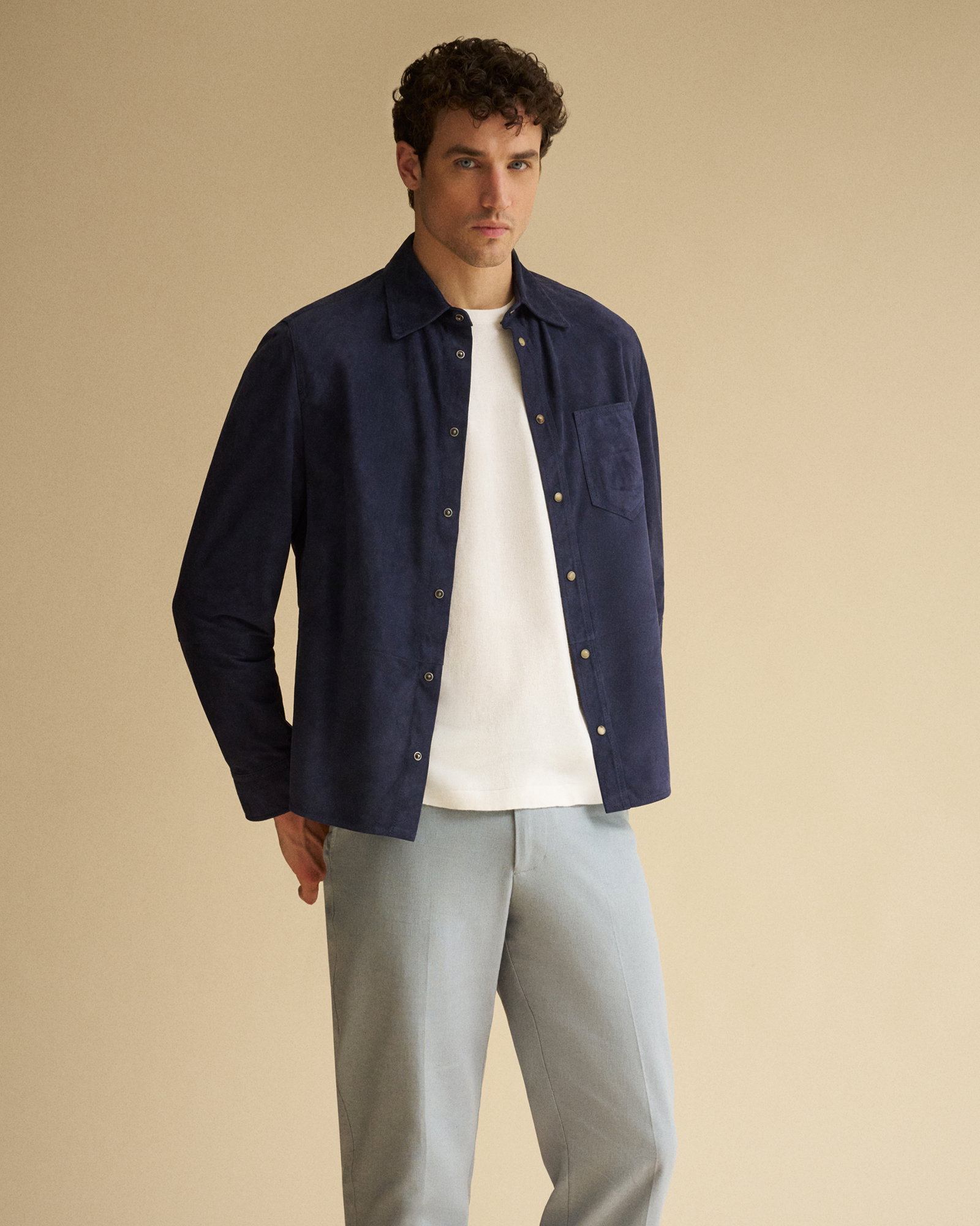 Mann mit lockigem Haar trägt dunkelblaues Overshirt, weißes T-Shirt und hellgraue Hose vor hellem Hintergrund.