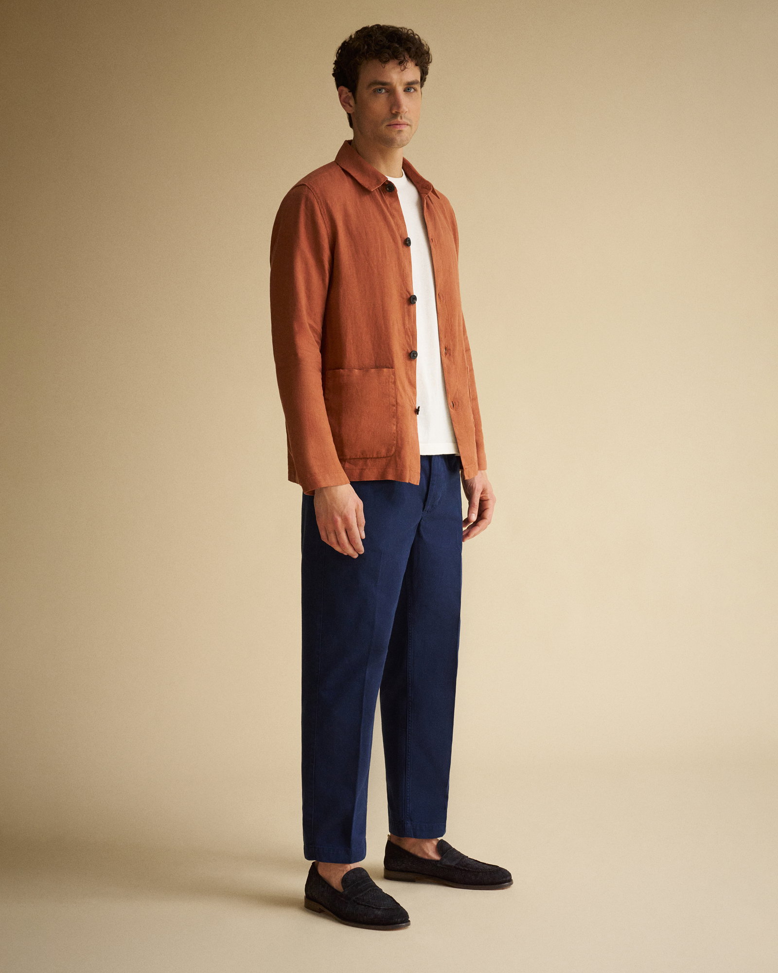 Mann trägt rostbraunes Overshirt, weißes T-Shirt, dunkelblaue Hose und schwarze Loafer, stehend vor beigem Hintergrund.