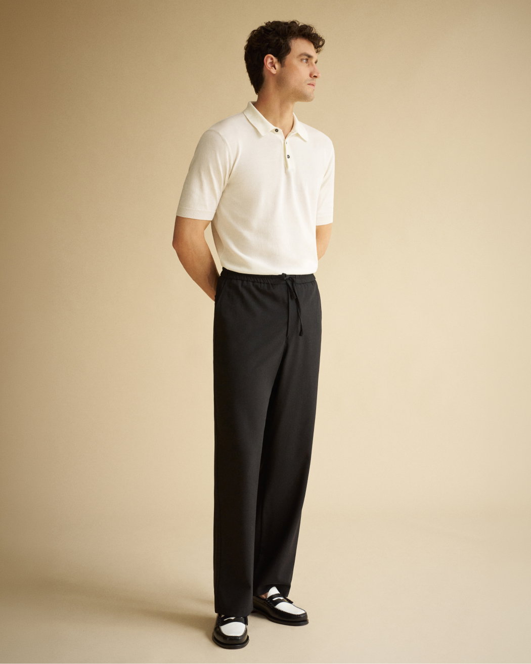 Mann trägt cremefarbenes Poloshirt, schwarze Hose mit Kordelzug und schwarz-weiße Loafer, steht vor hellem Hintergrund.