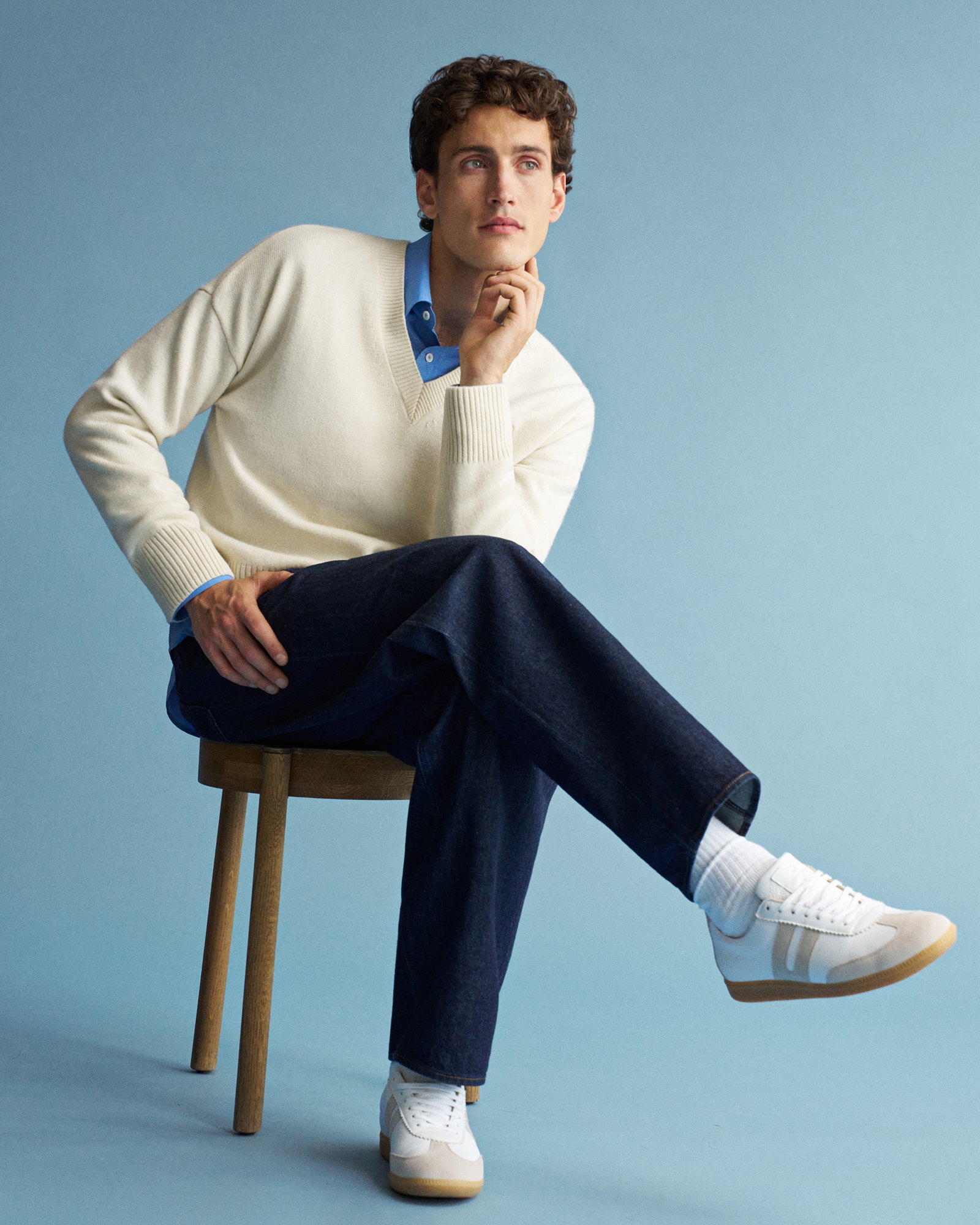 Mann sitzt auf Holzstuhl vor blauem Hintergrund, trägt cremefarbenen Pullover, hellblaues Hemd, dunkle Jeans und weiße Sneaker.