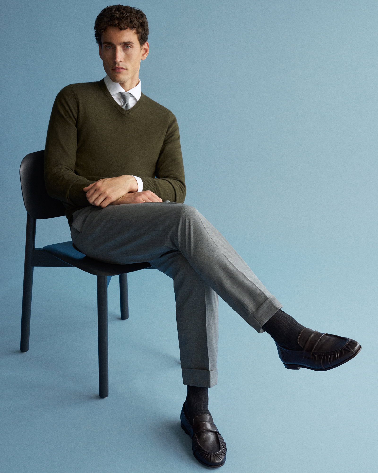 Mann sitzt auf Stuhl vor blauem Hintergrund, trägt olivgrünen Pullover, weißes Hemd mit Krawatte, graue Hose und braune Loafer.