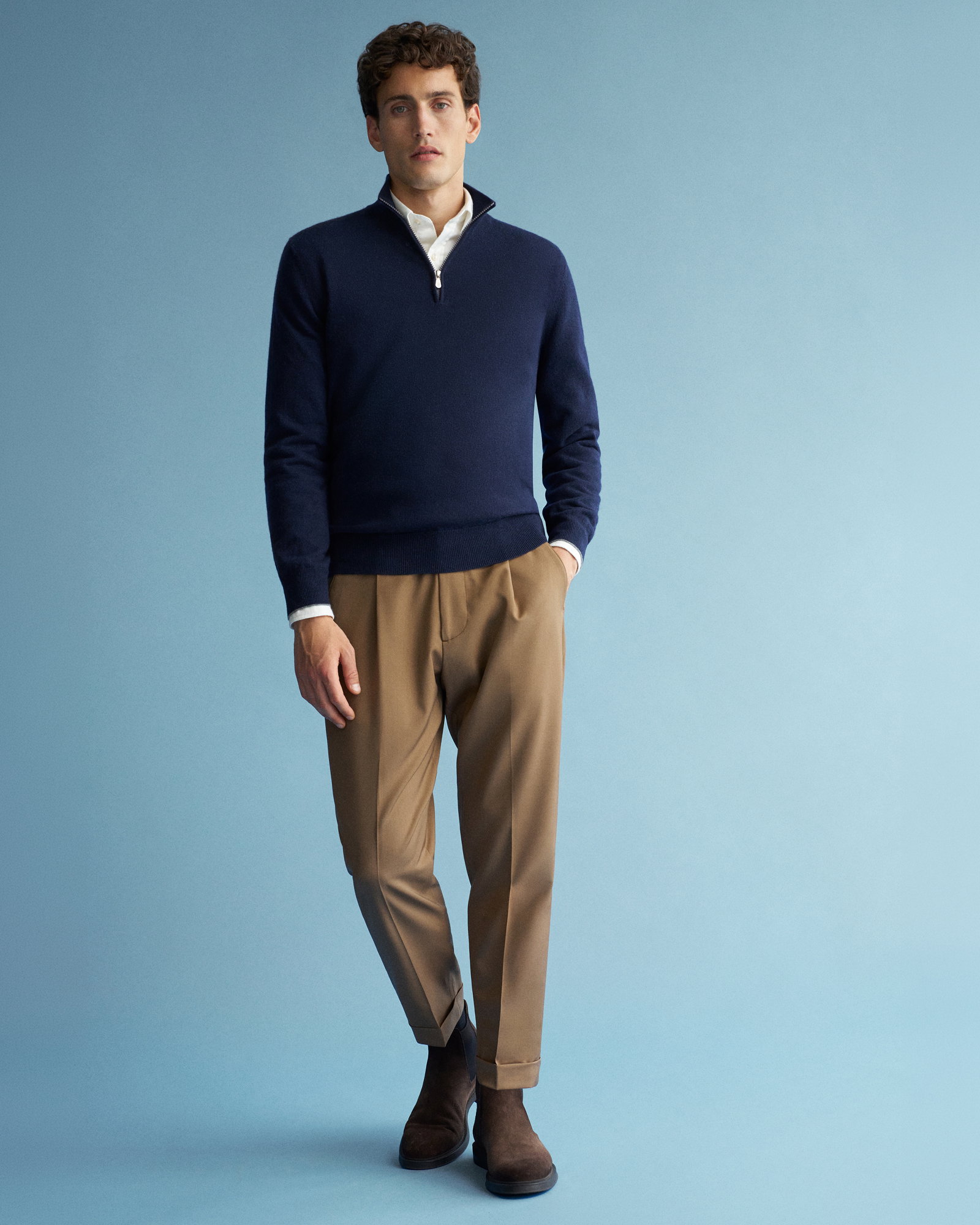 Mann trägt dunkelblauen Troyer mit Reißverschluss, weißes Hemd, hellbraune Hose und dunkelbraune Boots vor hellblauem Hintergrund.
