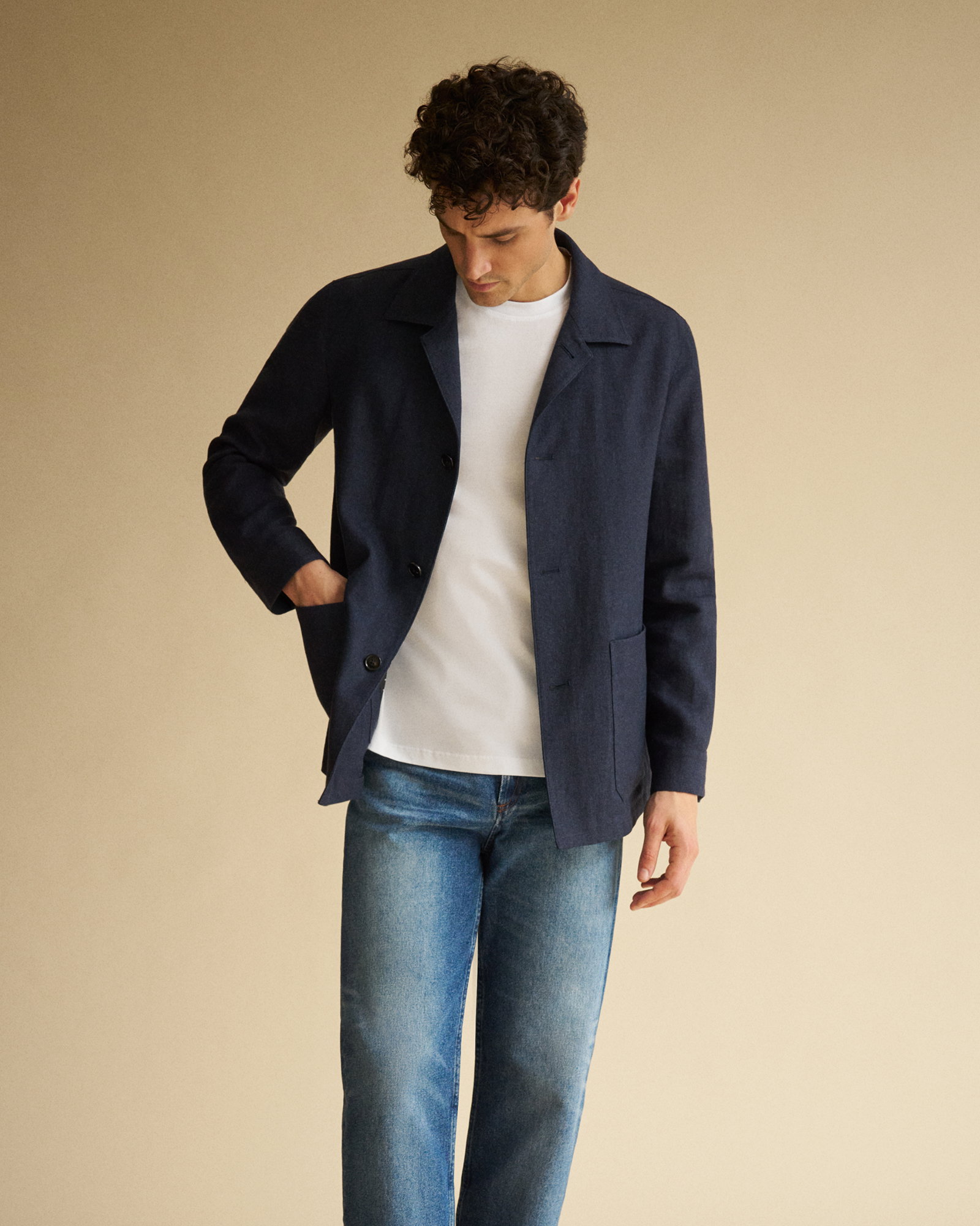 Mann mit dunklem Overshirt, weißem T-Shirt und blauen Jeans schaut nach unten vor hellem, einfarbigem Hintergrund.