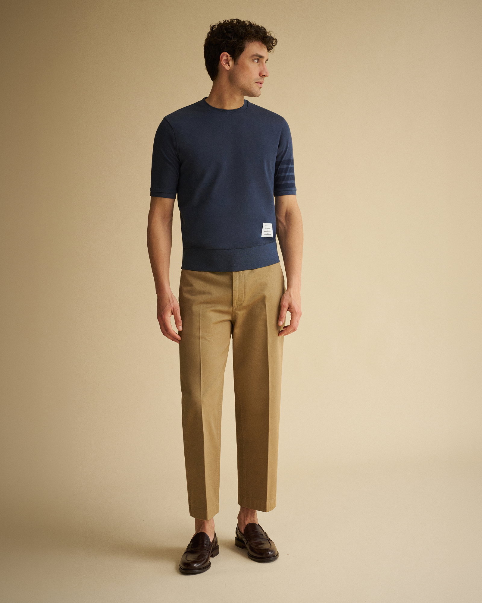 Mann trägt dunkelblaues T-Shirt mit Ärmelstreifen, beige Hose und dunkelbraune Loafer, steht vor hellem Hintergrund.