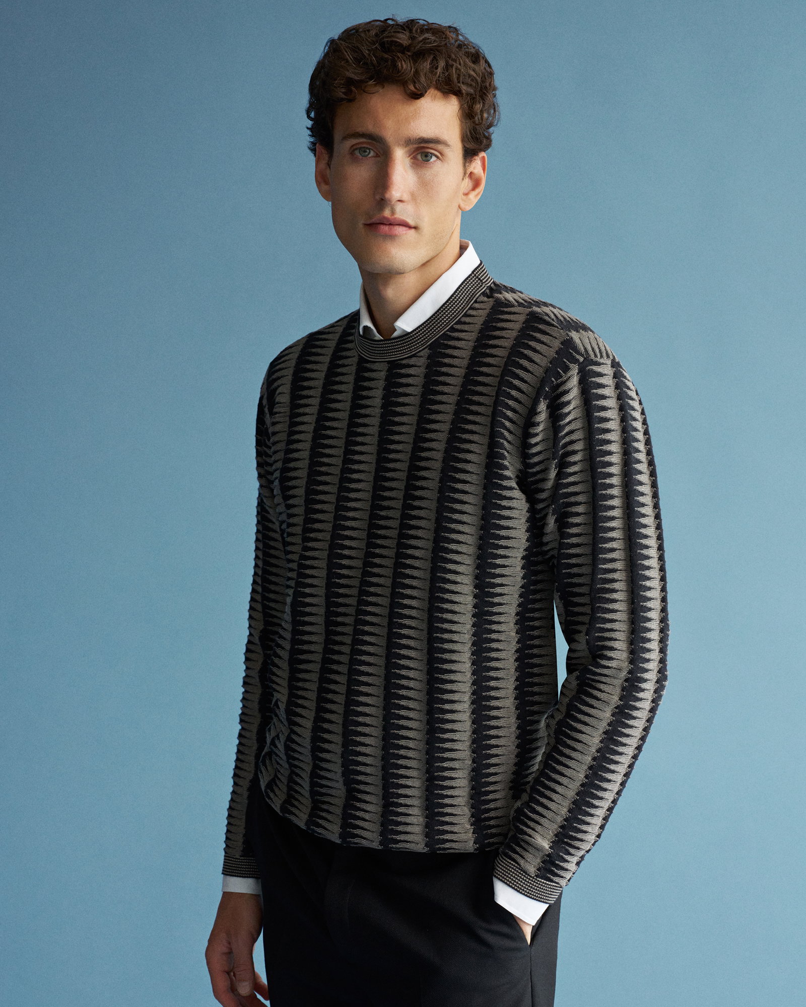 Junger Mann mit lockigem Haar trägt gemusterten schwarz-grauen Pullover über weißem Hemd vor blauem Hintergrund.