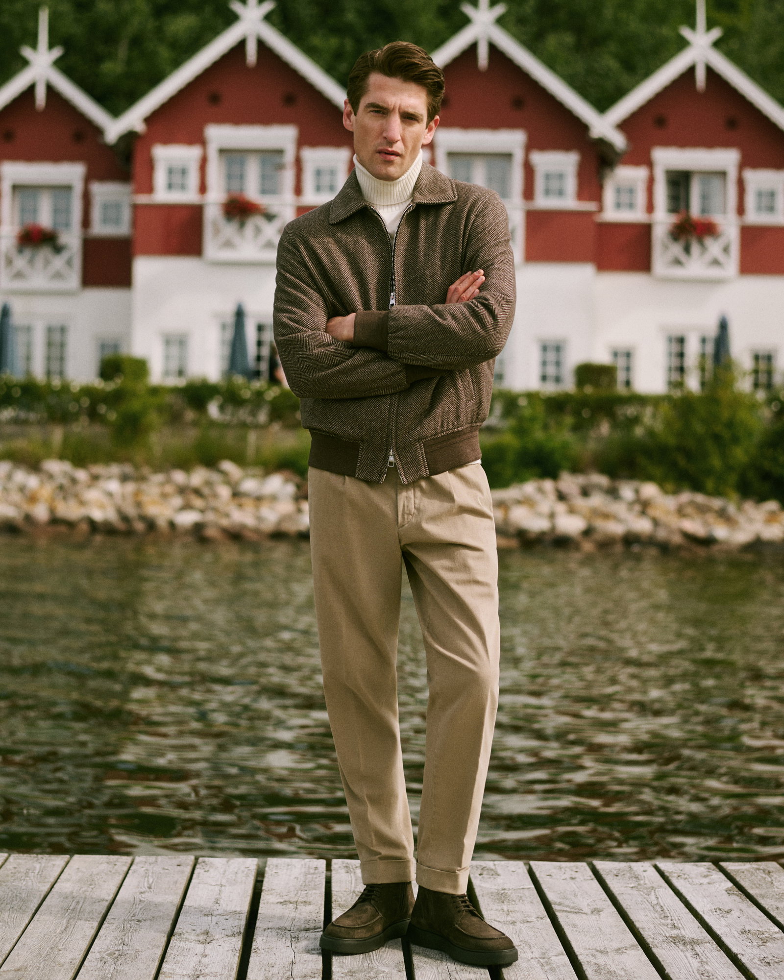 Mann steht mit verschränkten Armen auf Holzsteg, trägt braunen Blouson, cremefarbenen Rollkragenpullover, beige Hose vor skandinavischer Kulisse.