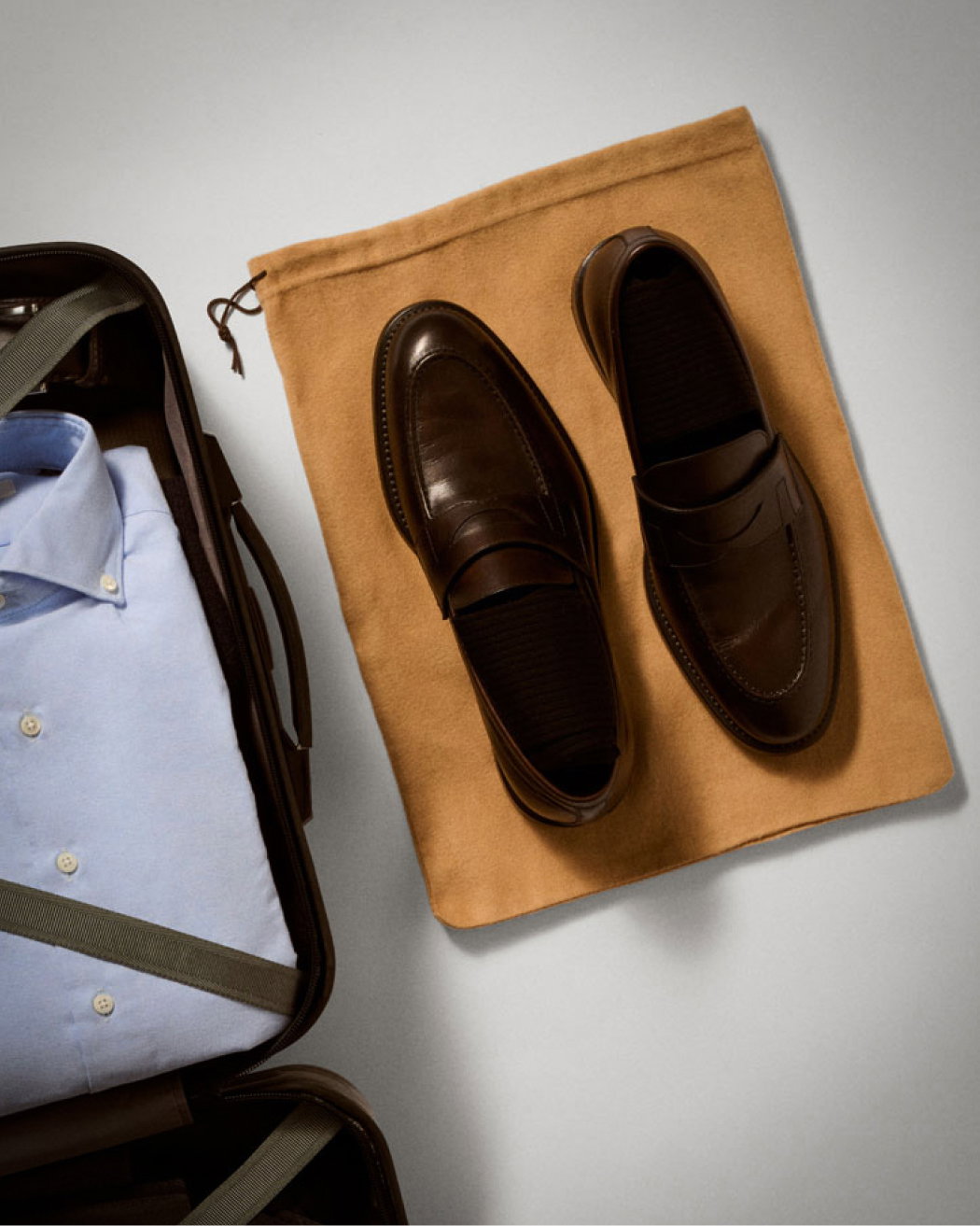 Braune Herren-Lederloafer auf beigem Staubbeutel neben offenem Koffer mit hellblauem Hemd und Gurten.