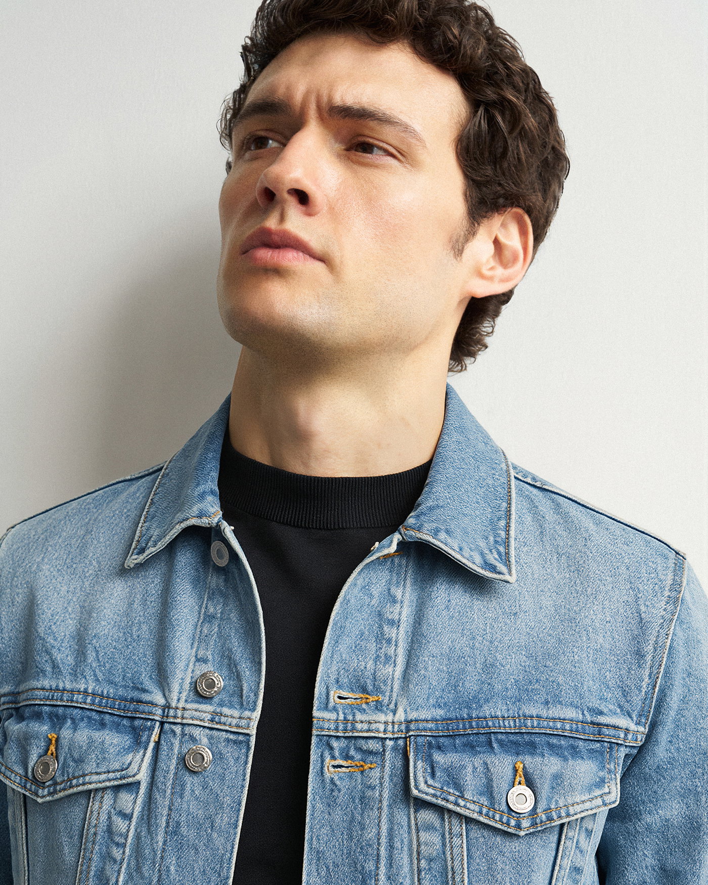 Porträt eines Mannes mit lockigem Haar, der ein schwarzes Shirt unter einer hellblauen Jeansjacke trägt, vor weißem Hintergrund.