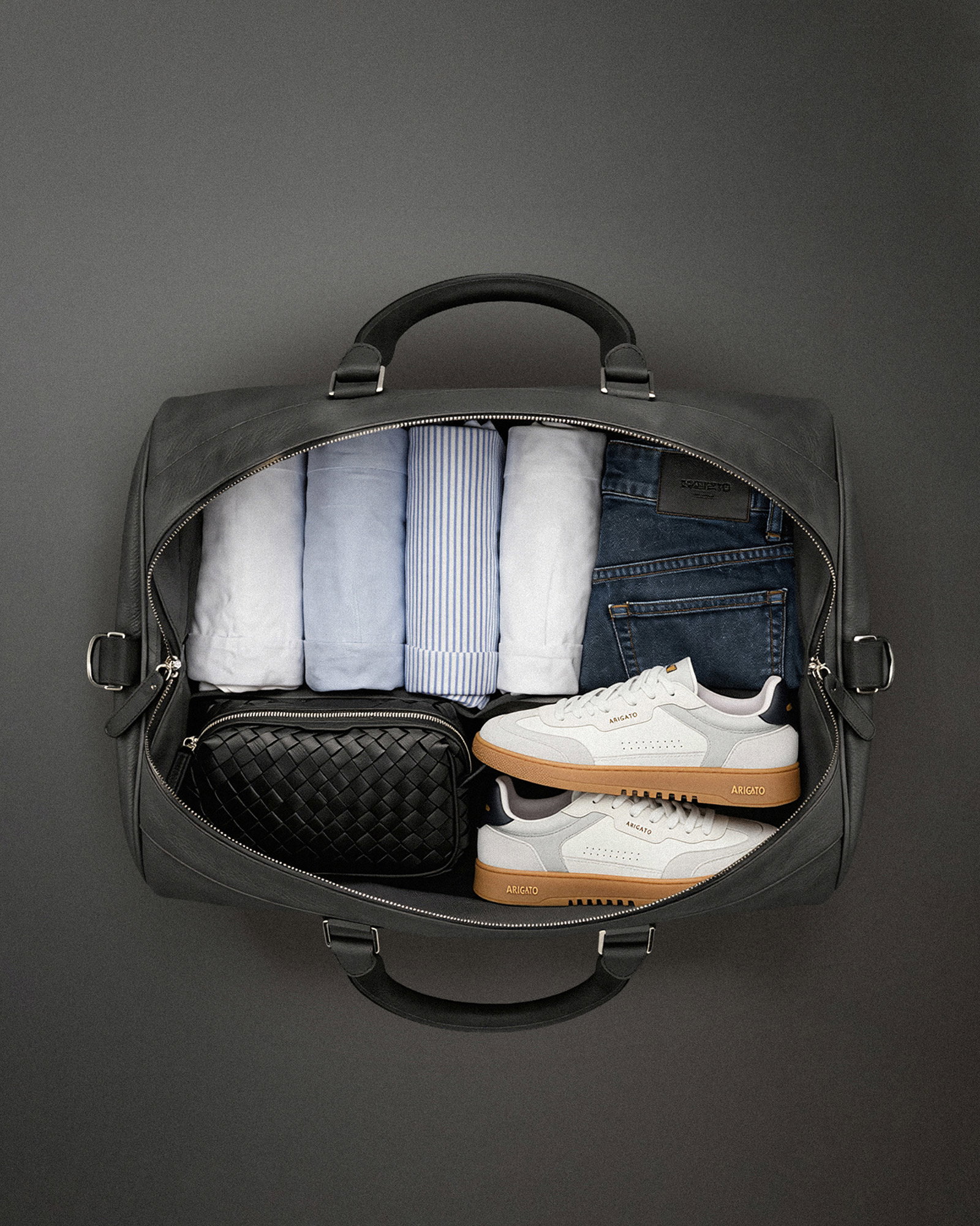 Offene schwarze Reisetasche mit fünf eingerollten Hemden, blauer Jeans, weißen Sneakern und schwarzem Necessaire.