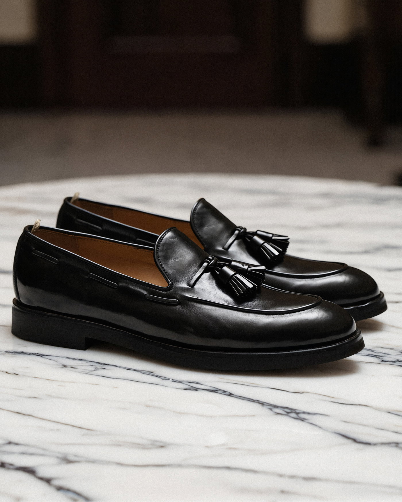 Schwarze Leder-Tasselloafer für Herren mit Quasten, sitzend auf weißem Marmoruntergrund, seitliche Ansicht.