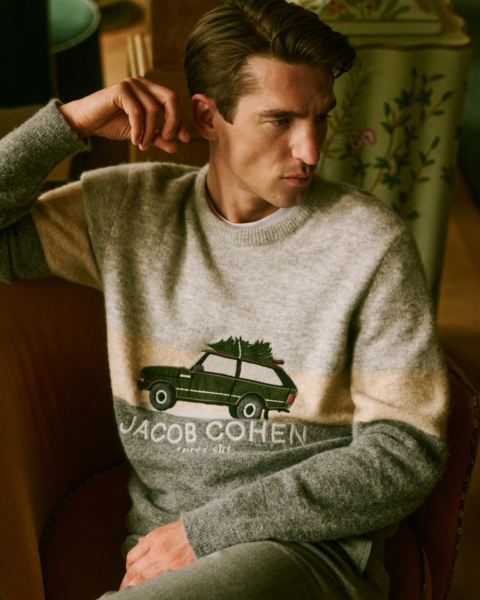 Sitzender Mann in grauem Rundhalspullover mit grünem Auto-Motiv und Schriftzug, beige-graue Hose, nachdenklich blickend.