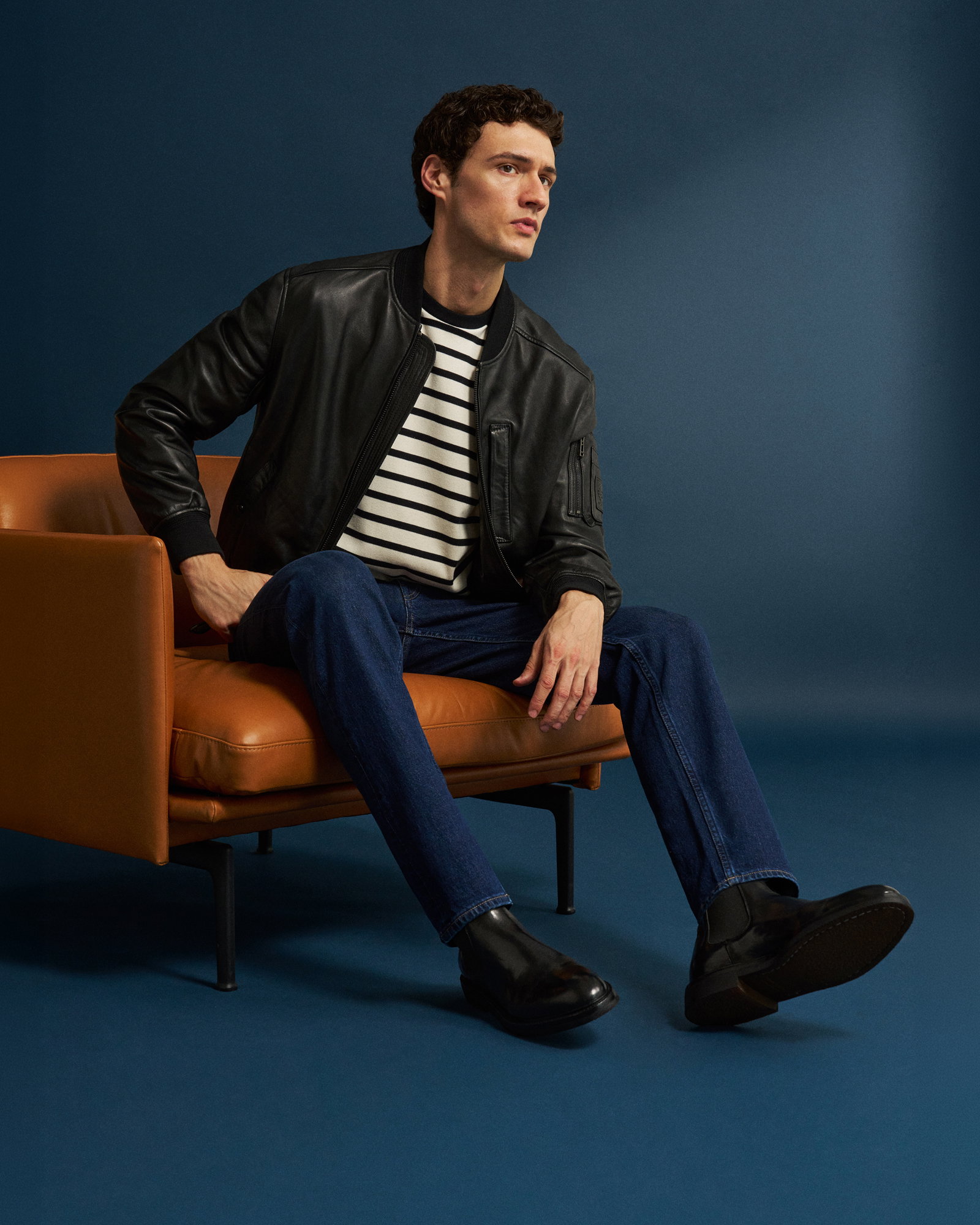 Sitzender Mann in schwarzer Lederjacke, gestreiftem T-Shirt, blauen Jeans und schwarzen Chelsea-Boots vor blauem Hintergrund.