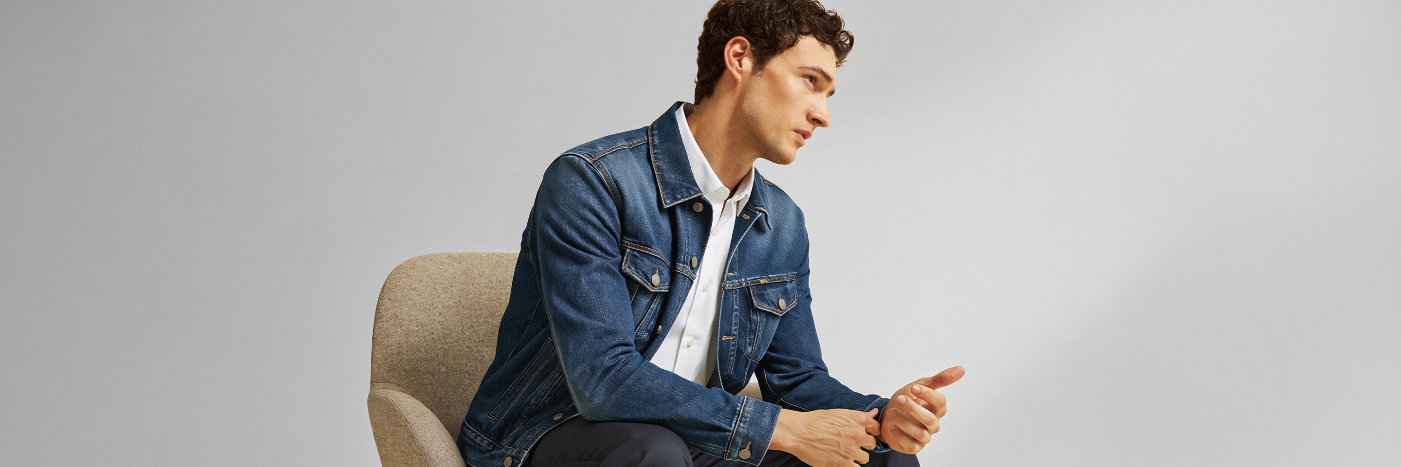 Sitzender Mann mit lockigem Haar, blauem Jeansjacke, weißem Hemd und dunkler Hose, schaut nach rechts, vor grauem Hintergrund.