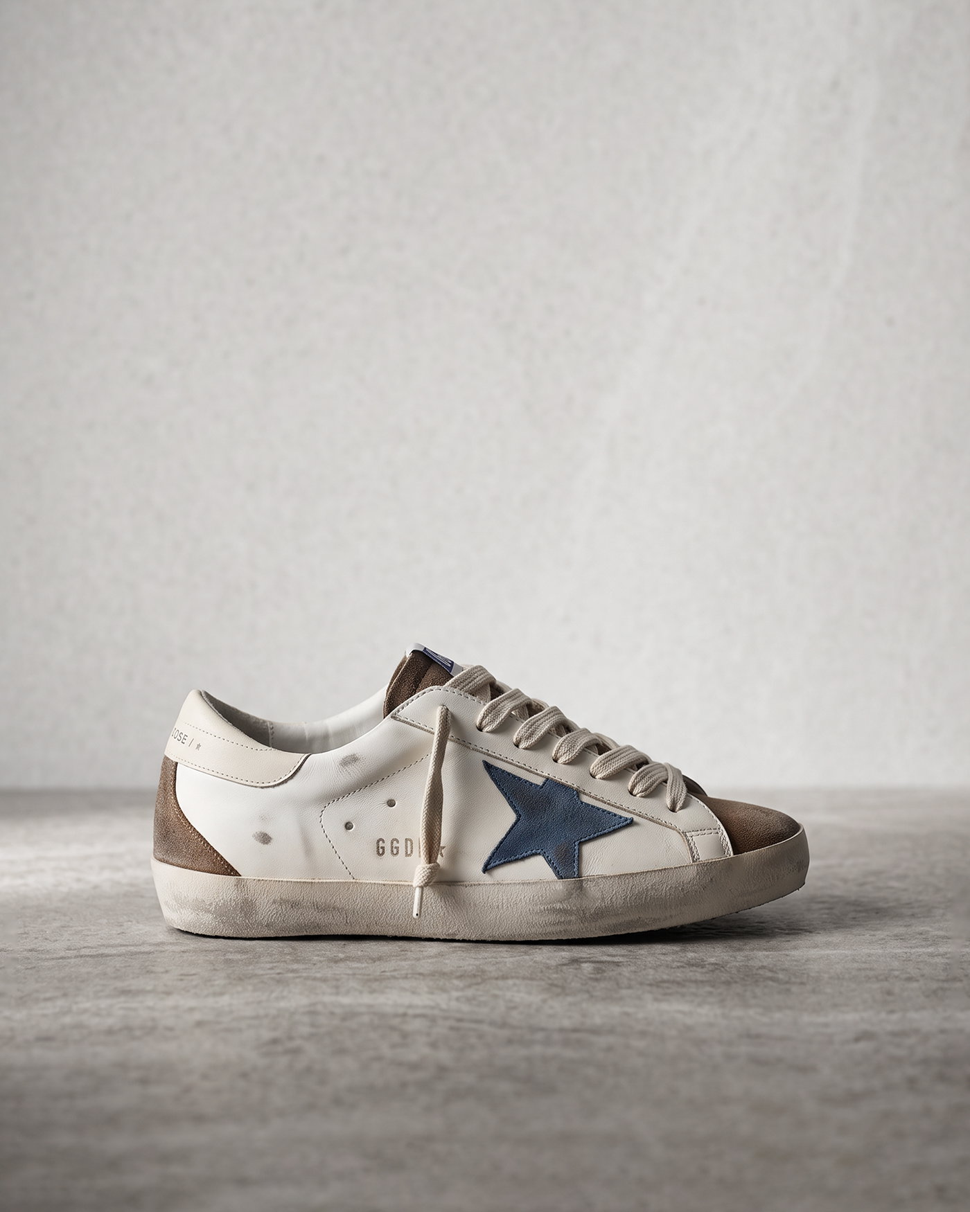Weiß-braune Herren-Sneaker im Used-Look mit blauem Stern und schmutzig wirkender Sohle auf grauem Steinboden.