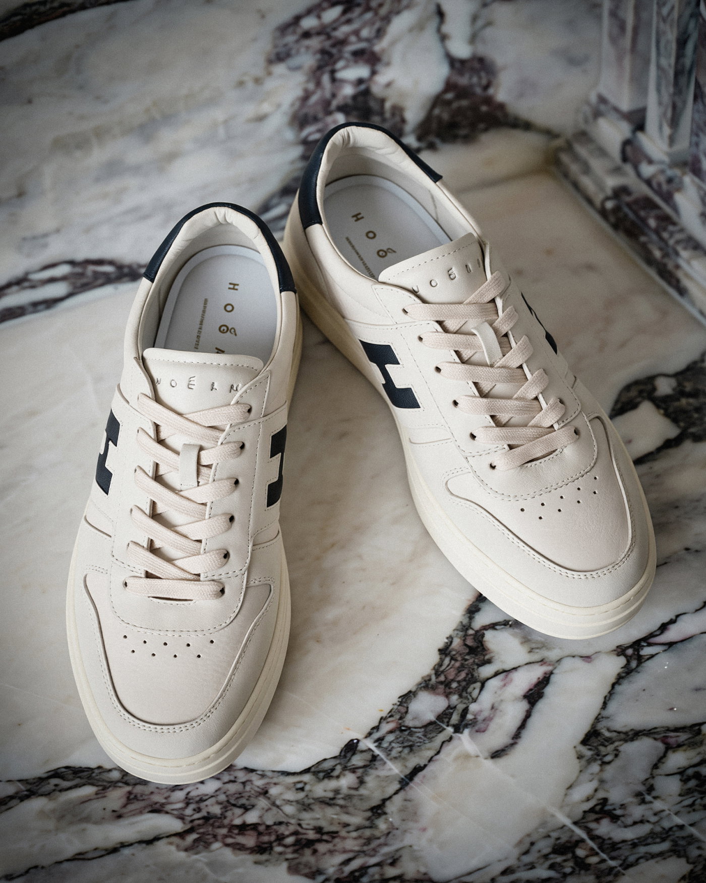 Weisse Herren-Sneaker mit schwarzem Designerlogo, beiger Sohle und Schnürsenkeln, auf gemustertem Marmorboden liegend.