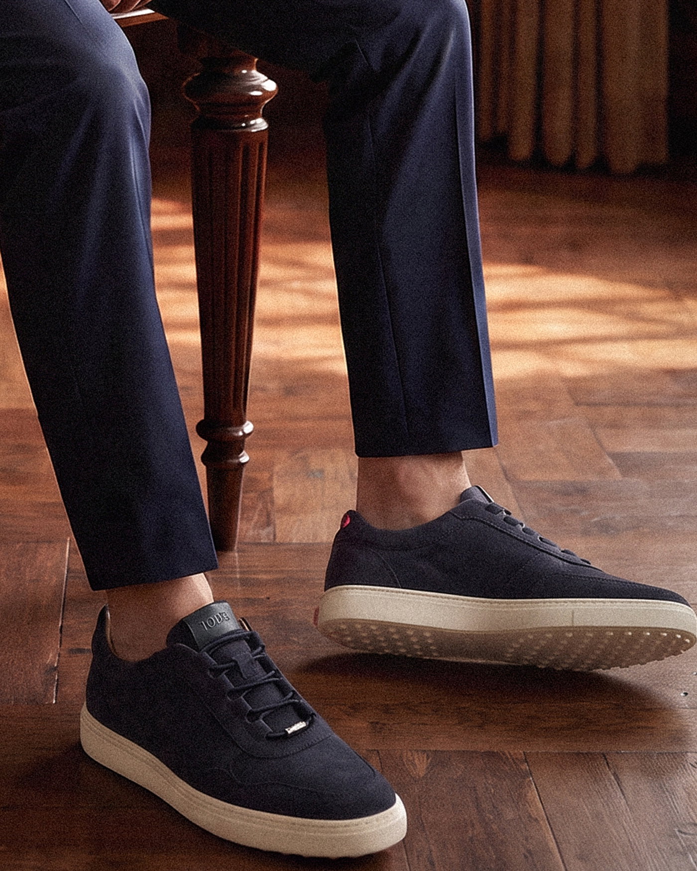 Wildledersneaker in Navy mit flexibler Schnürung, getragen zu dunkelblauer Herrenhose, auf braunem Holzboden sitzend.
