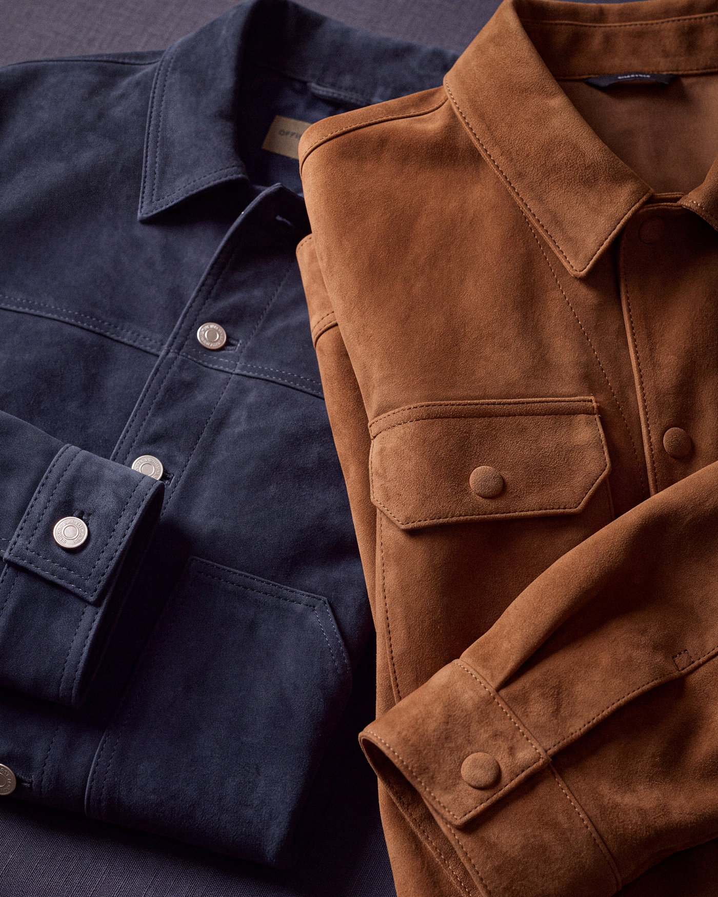 Zwei halb übereinandergelegte Herren-Velourslederjacken, eine in Navyblau und eine in Braun, mit Knöpfen und Taschen.