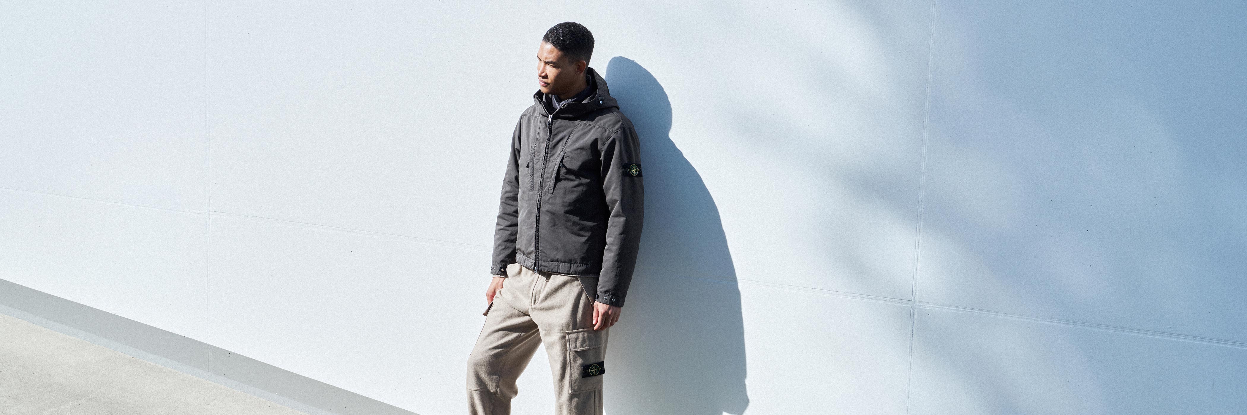 Junger Mann lehnt an weißer Wand, trägt schwarze Jacke mit Kapuze und beige Cargo-Hose, Schatten an der Wand sichtbar.
