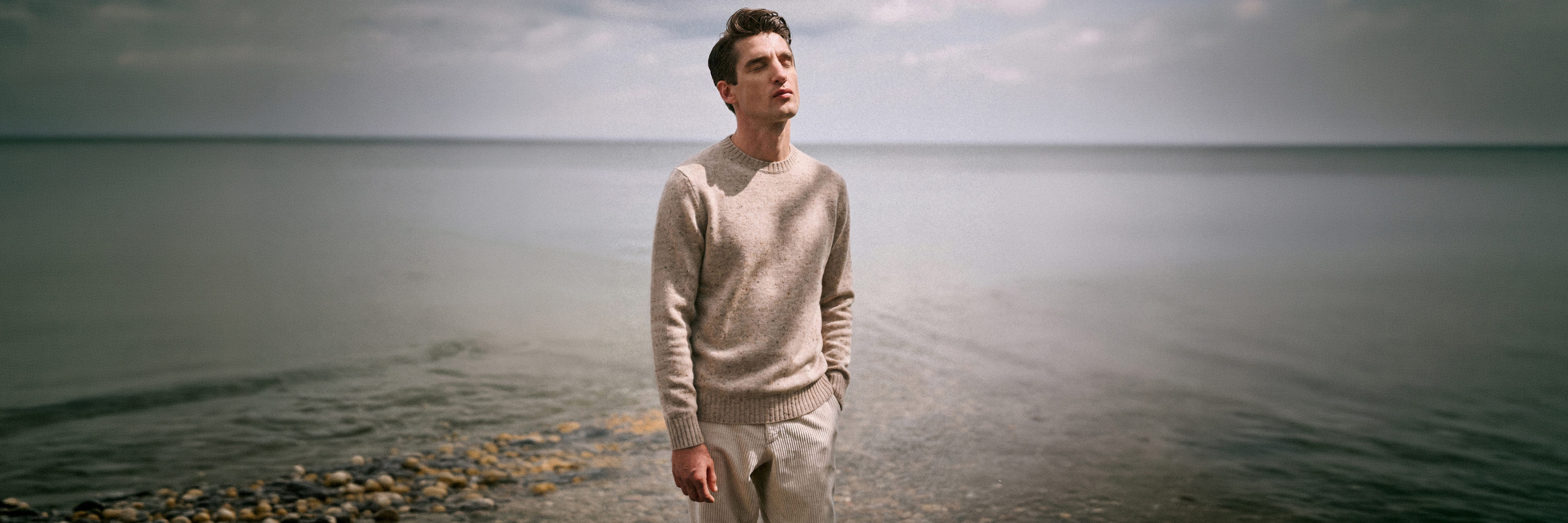 Junger Mann mit beigem Pullover und Hose steht entspannt am steinigen Ufer, Blick nach oben, ruhiges Meer im Hintergrund.