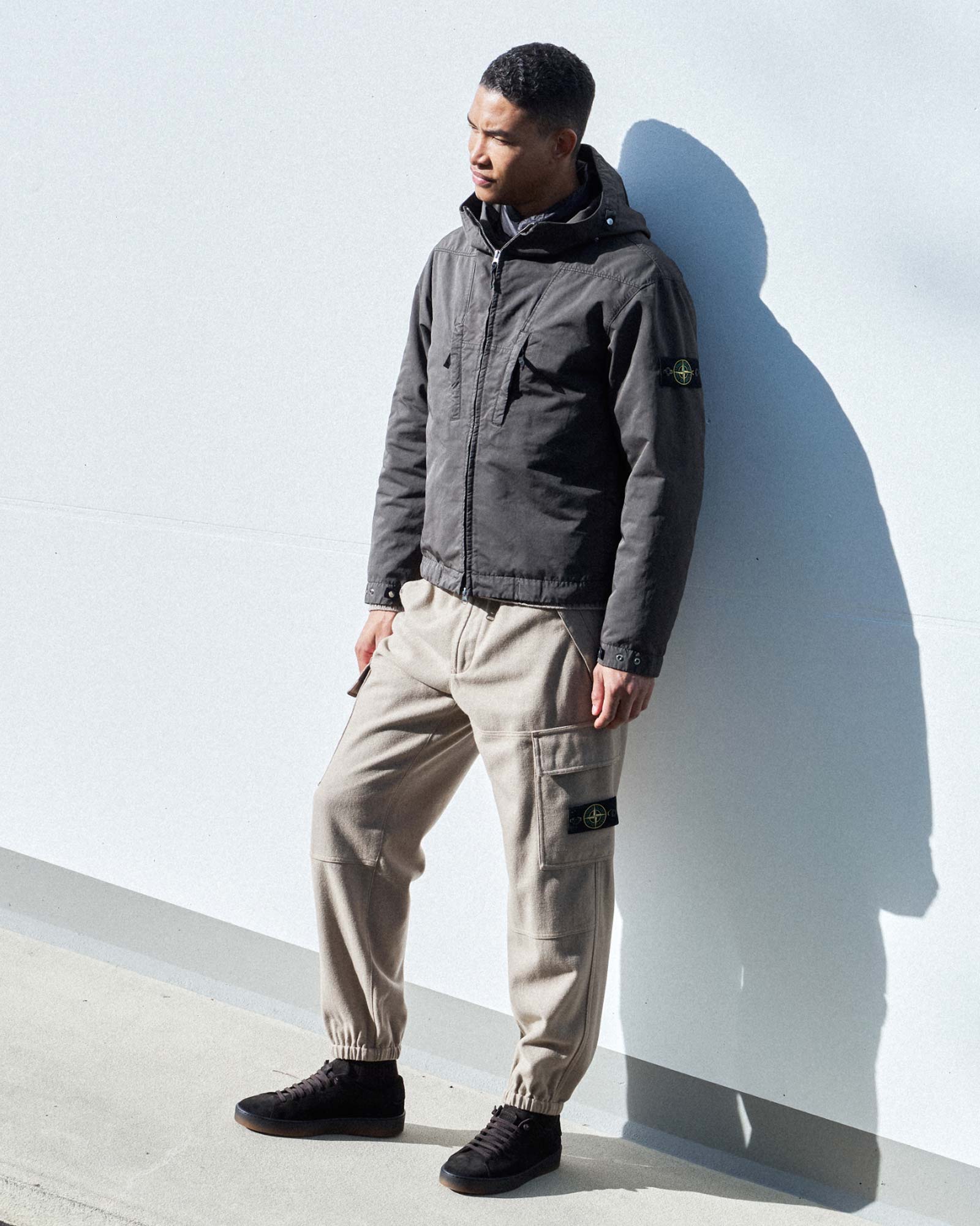 Mann lehnt an weißer Wand, trägt schwarze Jacke mit Kapuze, beige Cargo-Hose und schwarze Sneaker, Schatten an Wand.