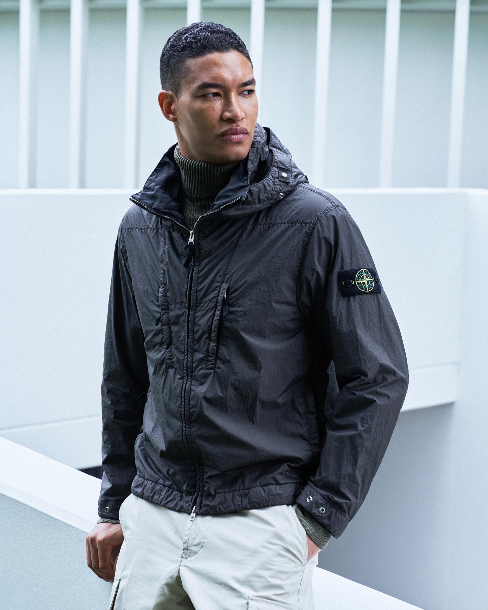 Mann mit kurzen lockigen Haaren trägt schwarze Jacke mit Kapuze und Stone Island-Logo, dazu helle Hose, vor weißem Hintergrund.