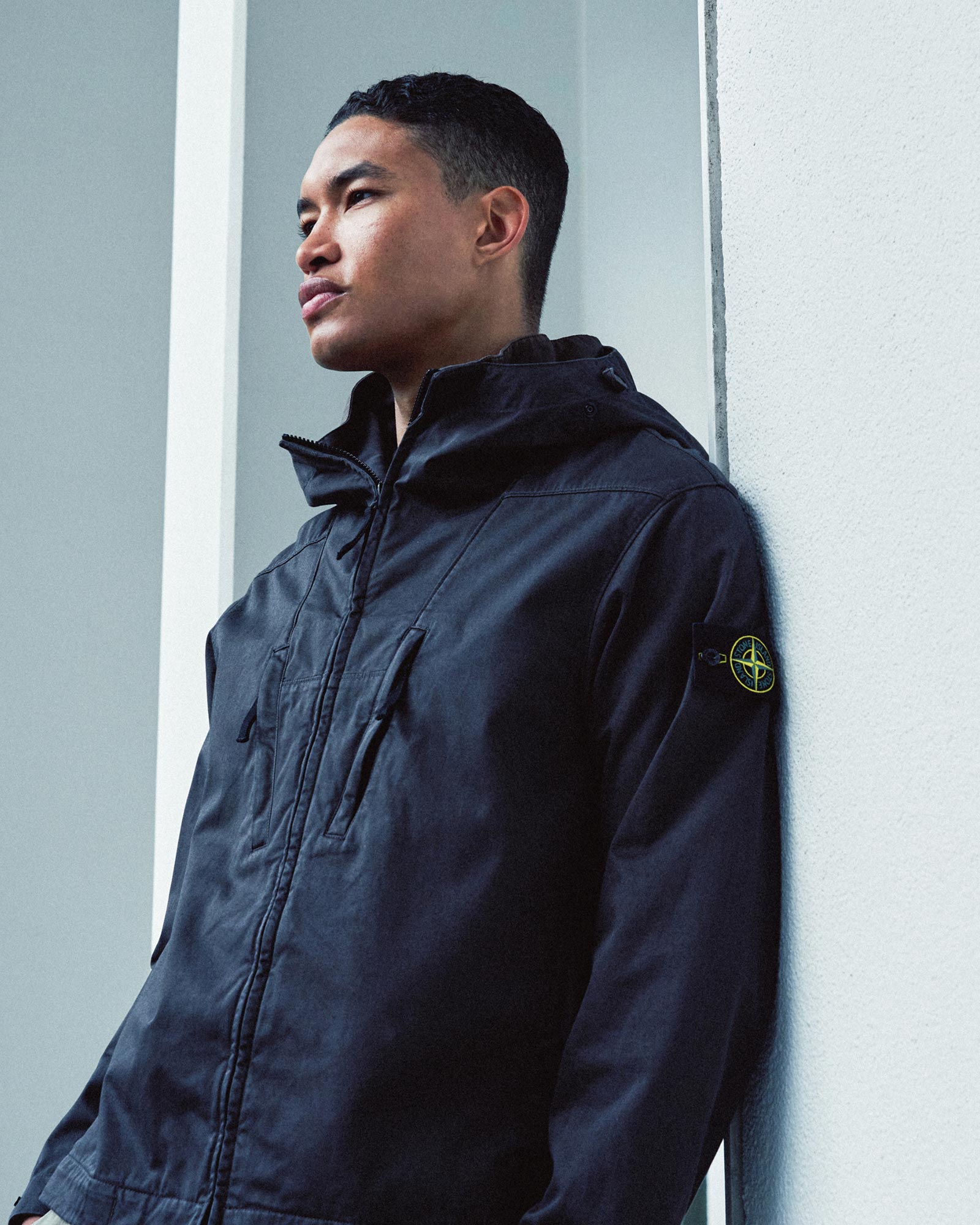 Junger Mann lehnt an weißer Wand, trägt schwarze Stone Island Jacke mit Logo am Ärmel, Blick nach links, urbaner Hintergrund.