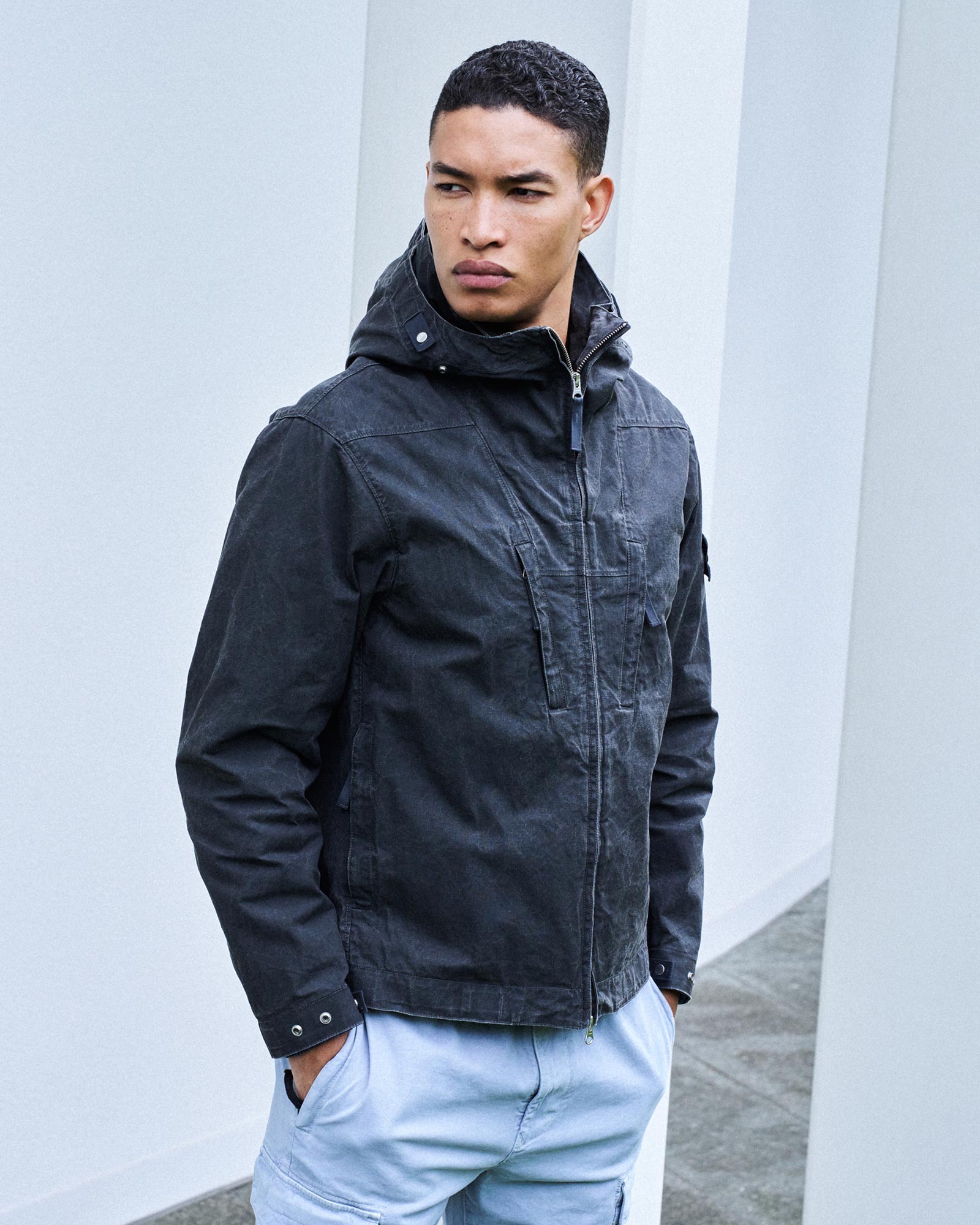 Junger Mann mit kurzen schwarzen Locken trägt schwarze Kapuzenjacke und hellblaue Hose vor weißer Wand.