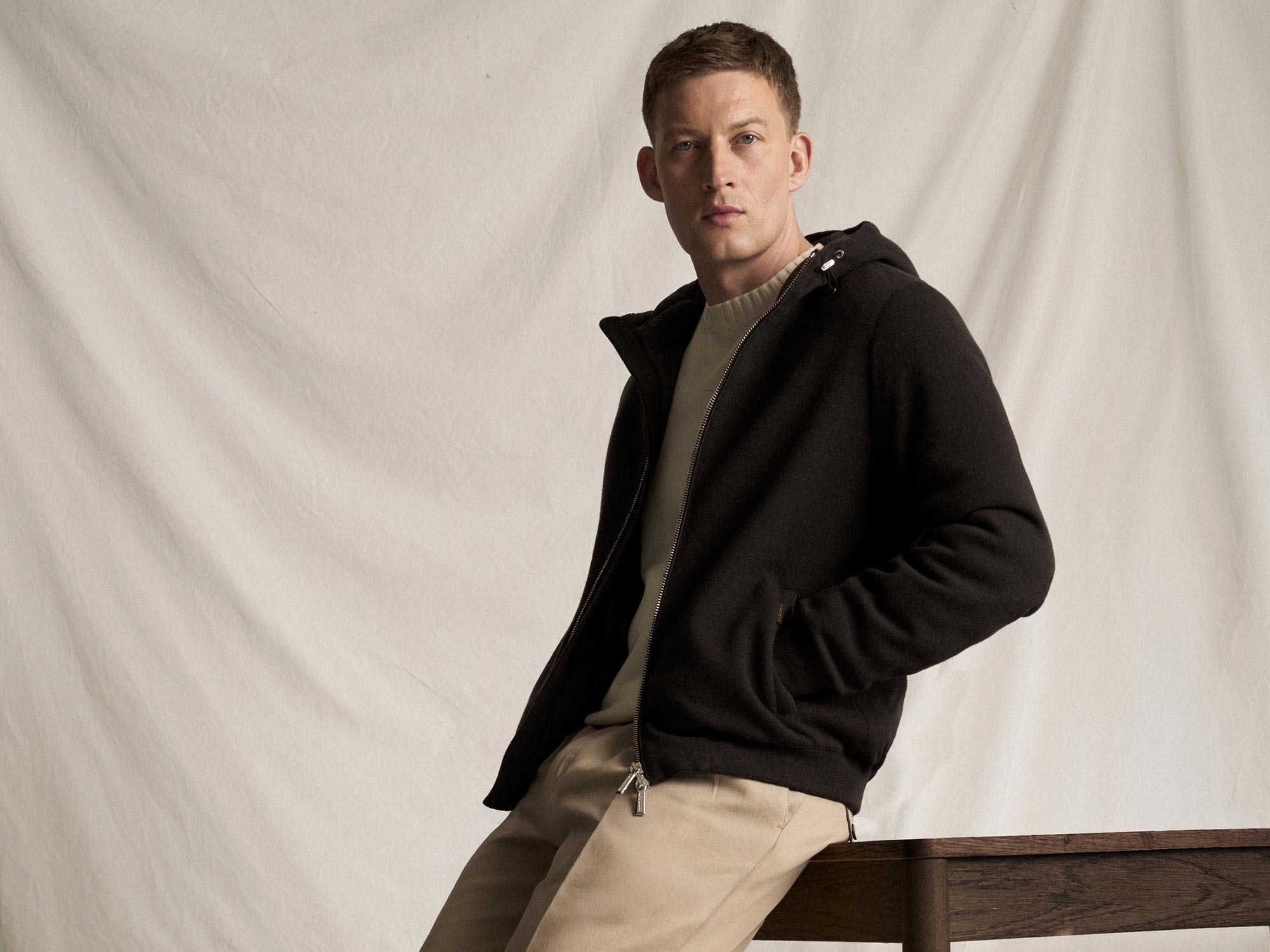 Junger Mann mit kurzem braunem Haar trägt schwarzen Hoodie, hellen Pullover und beige Hose, sitzt vor hellem Hintergrund.