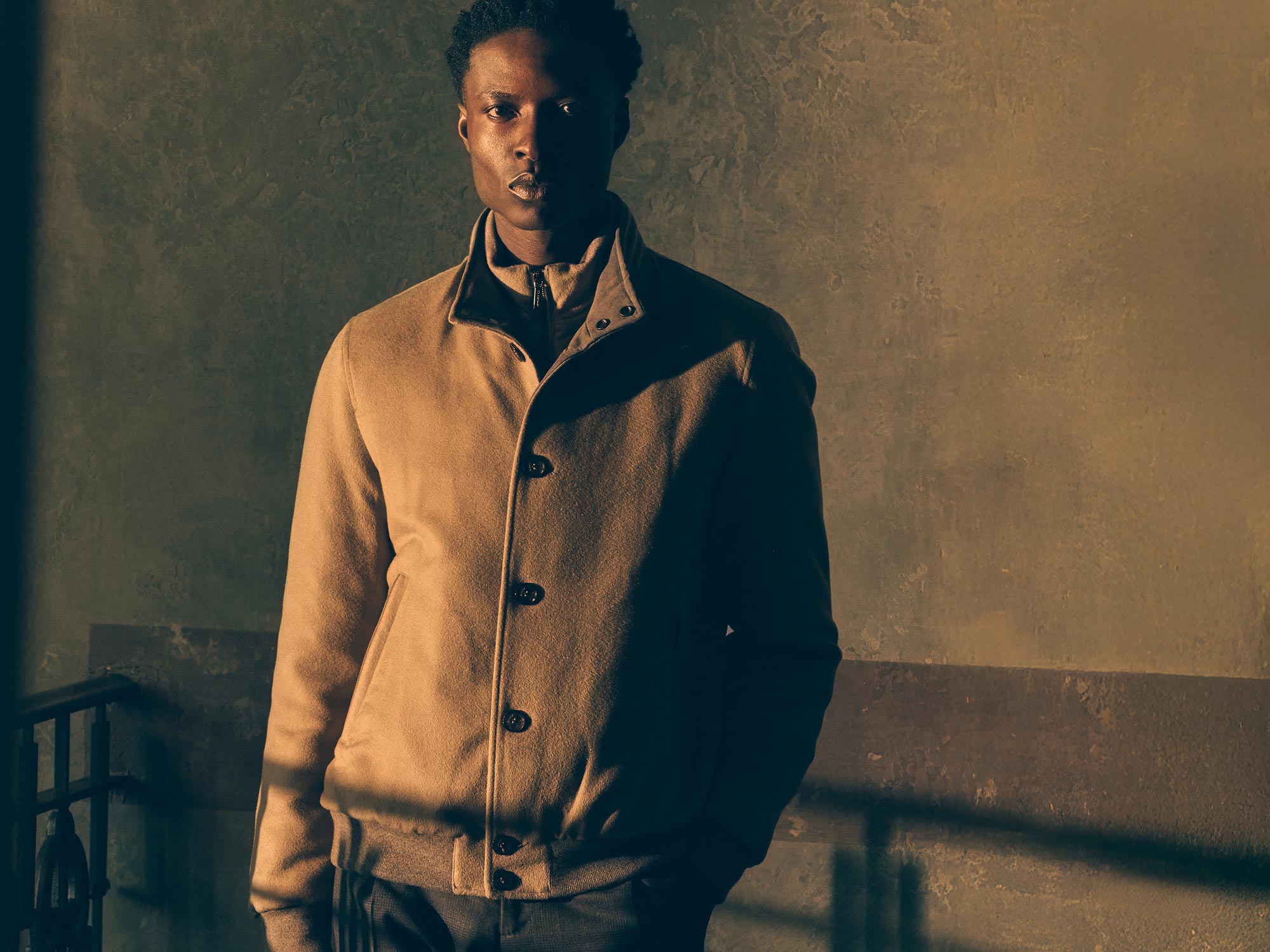 Young man wearing a beige buttoned coat standing against a textured wall with dramatic lighting casting shadows.