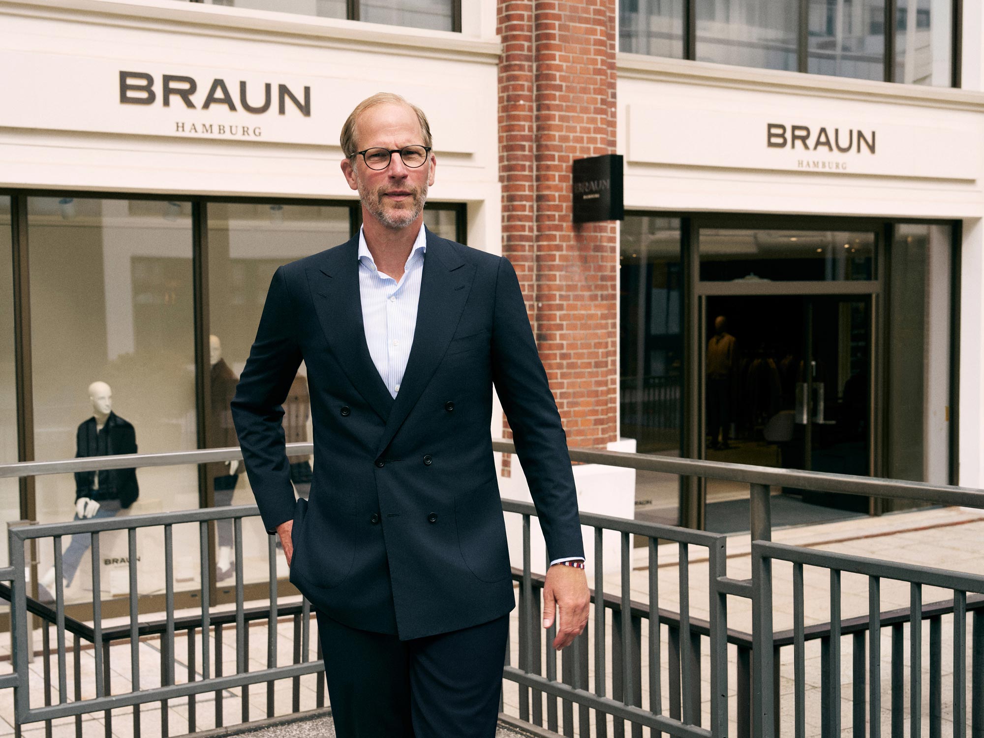 Mann mit Brille und Anzug steht vor dem Geschäft BRAUN Hamburg, im Hintergrund Schaufenster mit Mode und roter Backsteinwand.