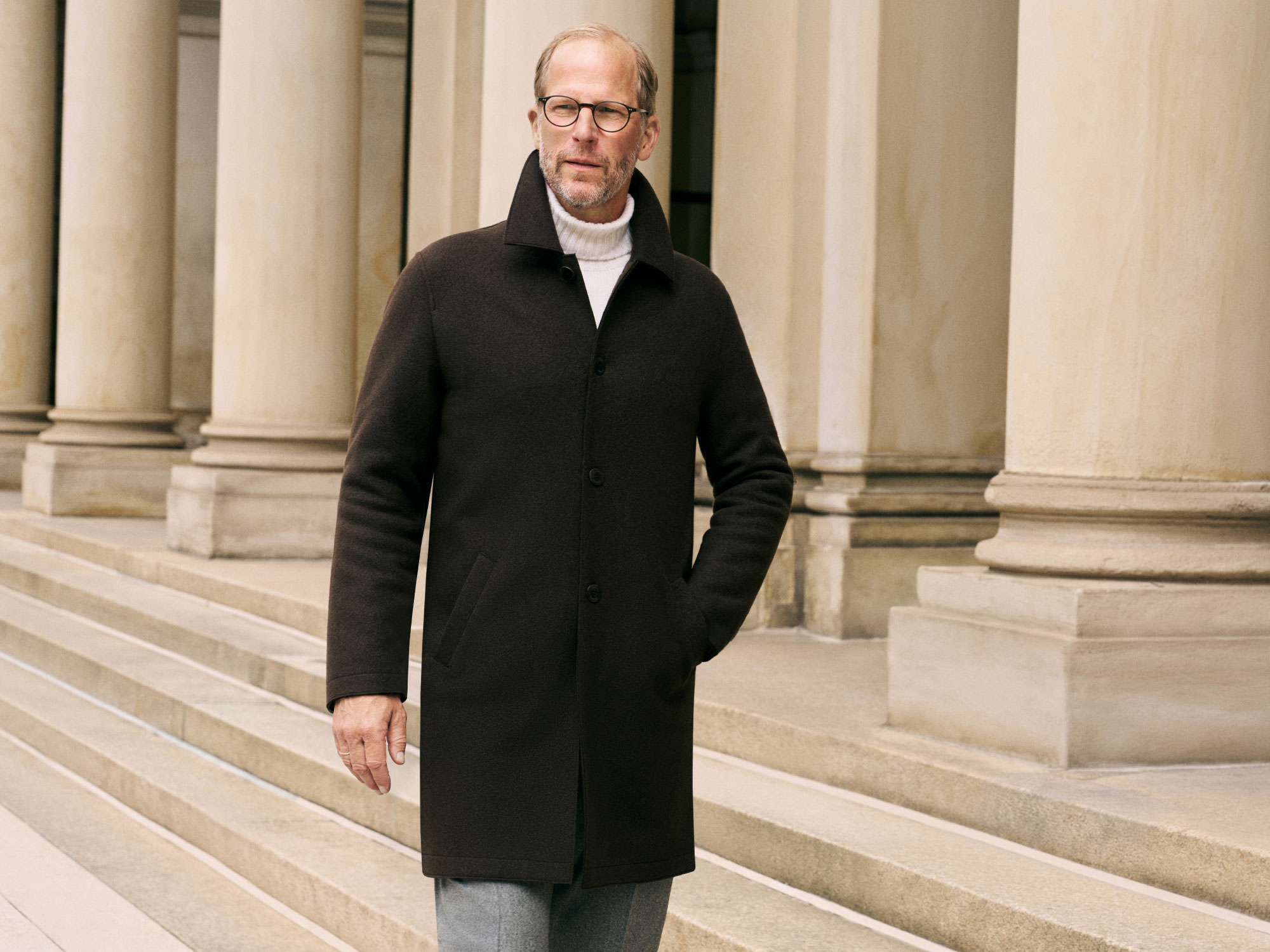 Man wearing round glasses, dark brown coat, white turtleneck, and gray pants standing on stone steps by large beige columns.