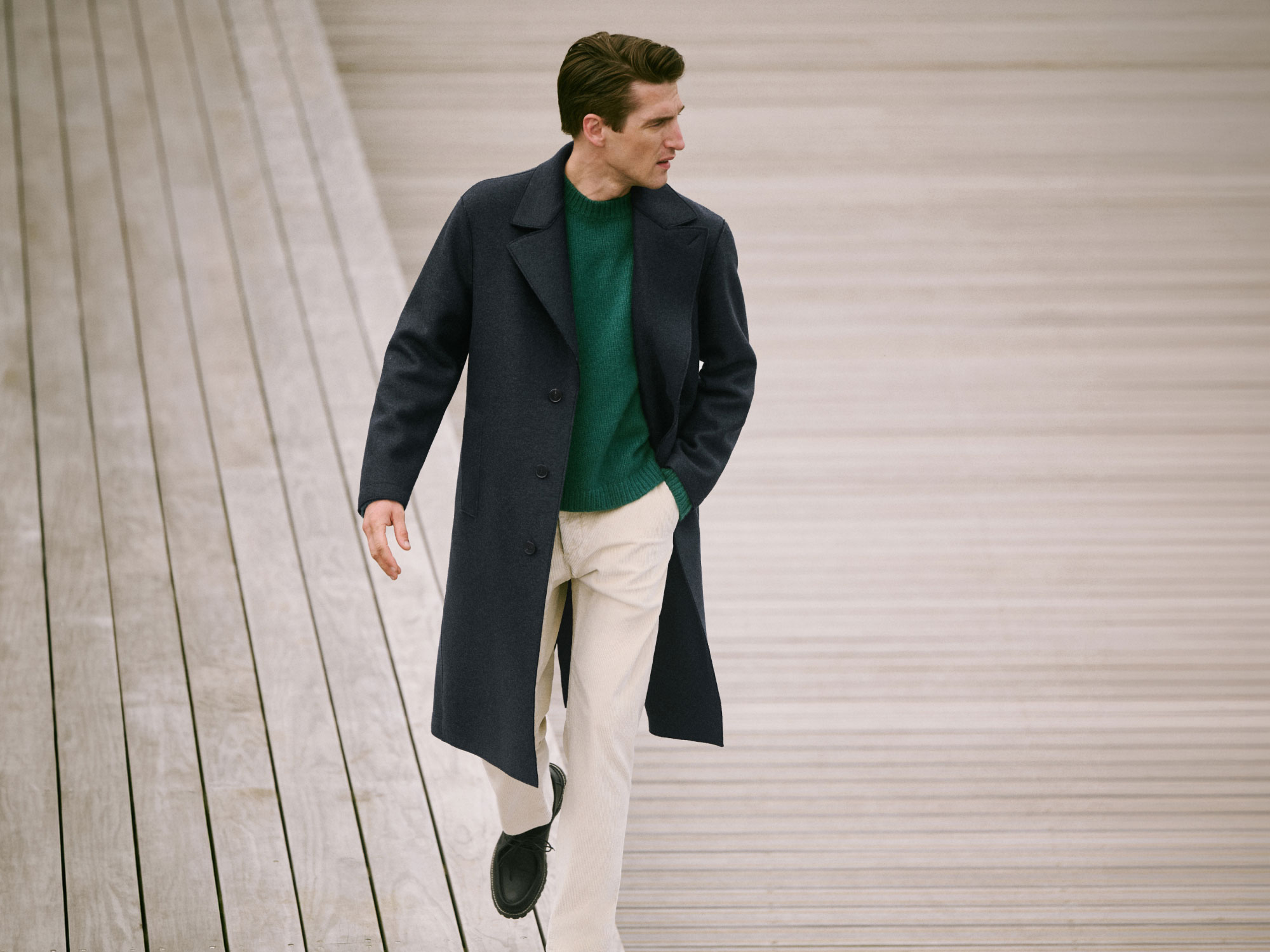 Man walking on wooden stairs wearing a dark long coat, green sweater, beige pants, and black shoes, looking to the side.