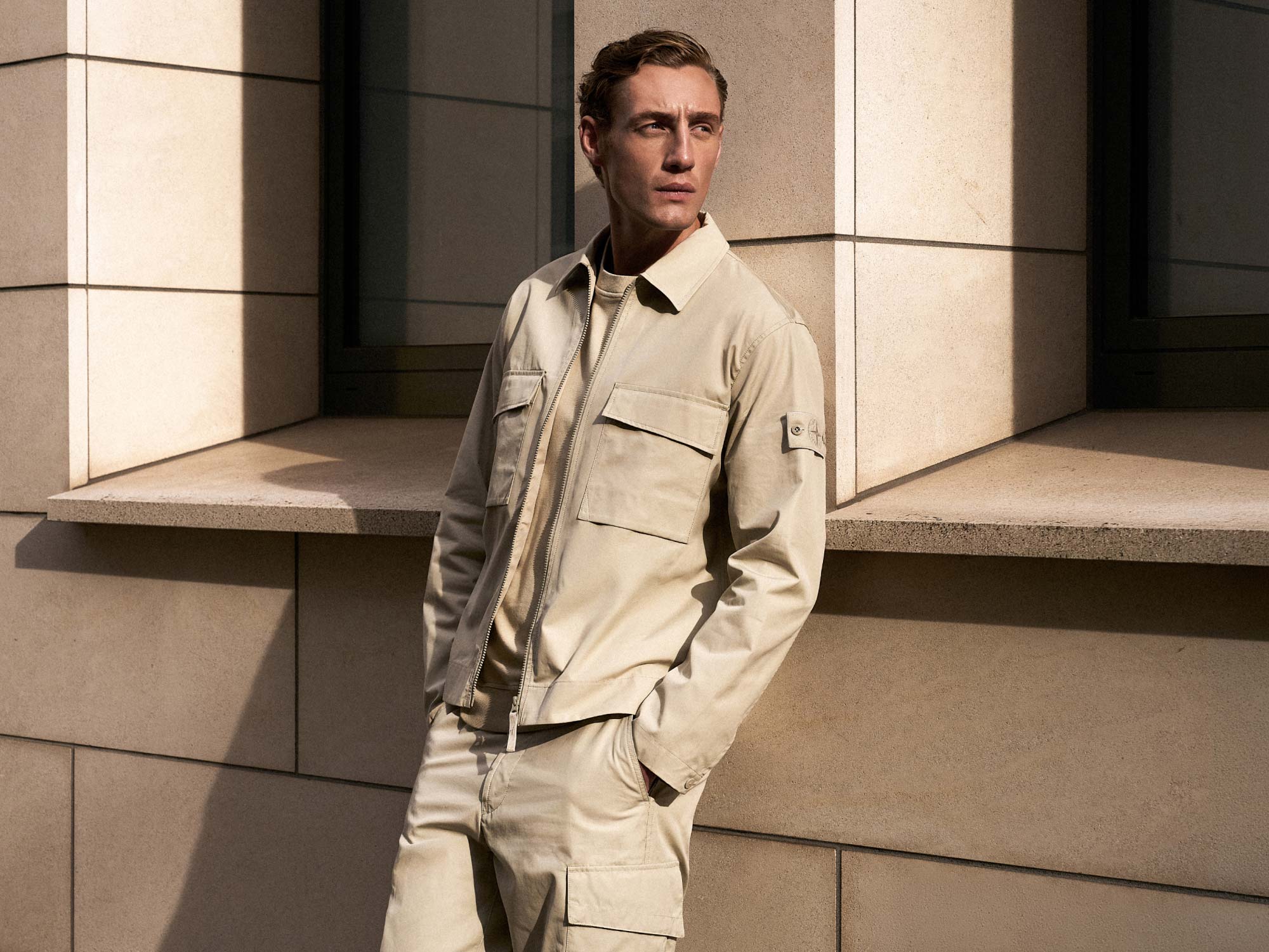 Mann lehnt an hellem Steinmauer-Eck, trägt beige Jacke mit Taschen und Hose, schaut nach rechts in Sonnenlicht.